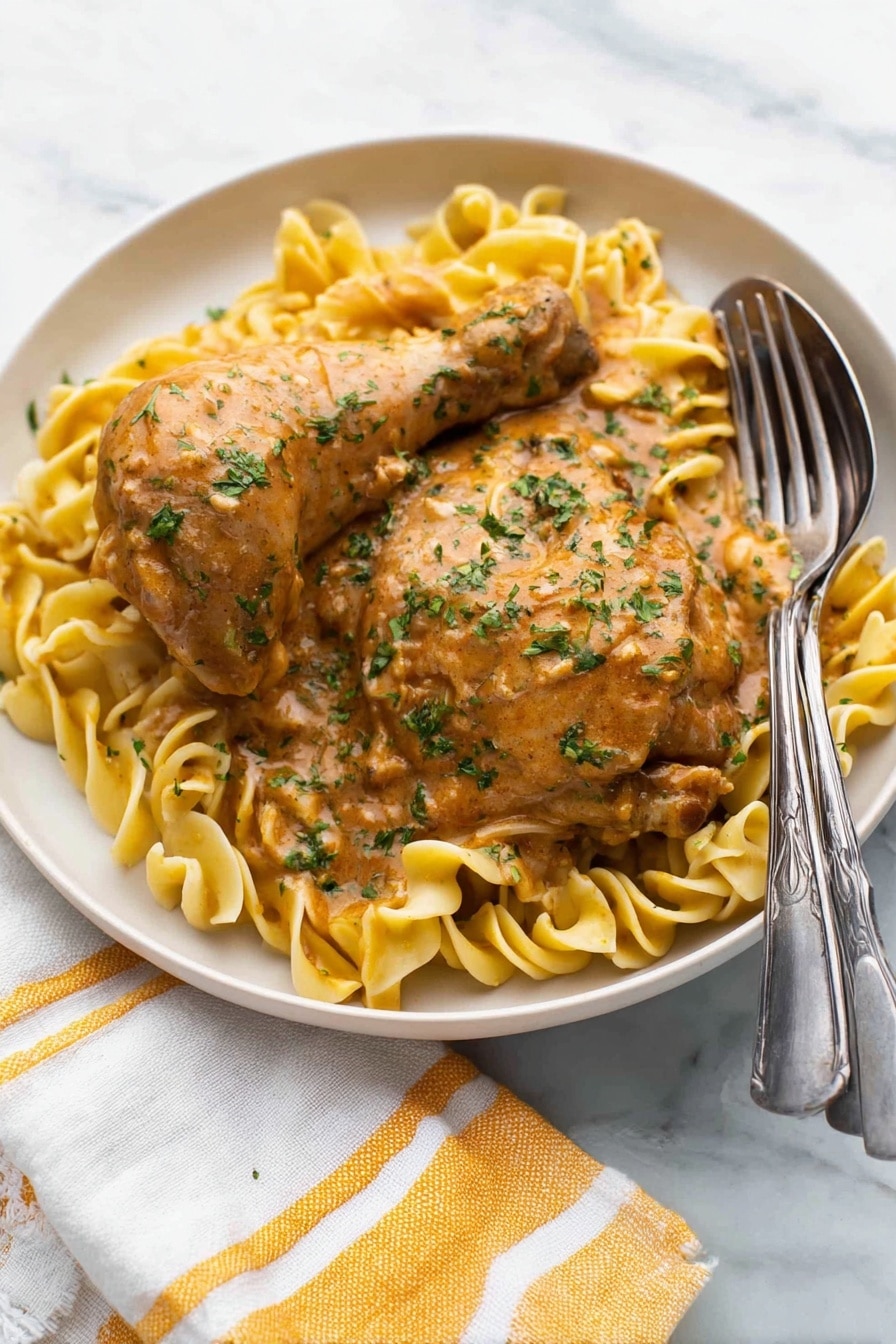 A white round plate holds two pieces of cooked chicken, one drumstick and one thigh, both covered in a thick light brown sauce with green herbs sprinkled on top. Underneath, there is a bed of light yellow egg noodles twisted softly, also coated lightly with the same sauce. A silver fork and spoon rest on the right edge of the plate. The plate sits on a white marbled surface with a white and yellow striped cloth napkin partially visible at the bottom left corner. Photo taken with an iphone --ar 2:3 --v 7 - Easy Chicken Paprikash, Hungarian chicken dish, creamy paprika chicken, slow-cooker paprikash, traditional Hungarian recipes