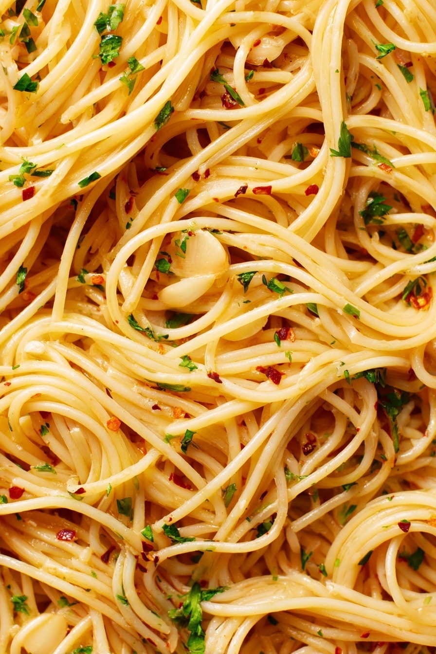 The image shows a close-up view of spaghetti pasta with thin, long noodles that are light golden in color. The noodles are mixed evenly with thin slices of light tan garlic, small bits of green parsley, and tiny red chili flakes scattered throughout. The spaghetti appears glossy with a slightly oily texture, making the noodles look smooth and shiny. The background is a white marbled texture. photo taken with an iphone --ar 2:3 --v 7 - Garlic Spaghetti Aglio e Olio, easy garlic pasta recipe, quick spaghetti dinner, simple Italian pasta dish, flavorful garlic pasta