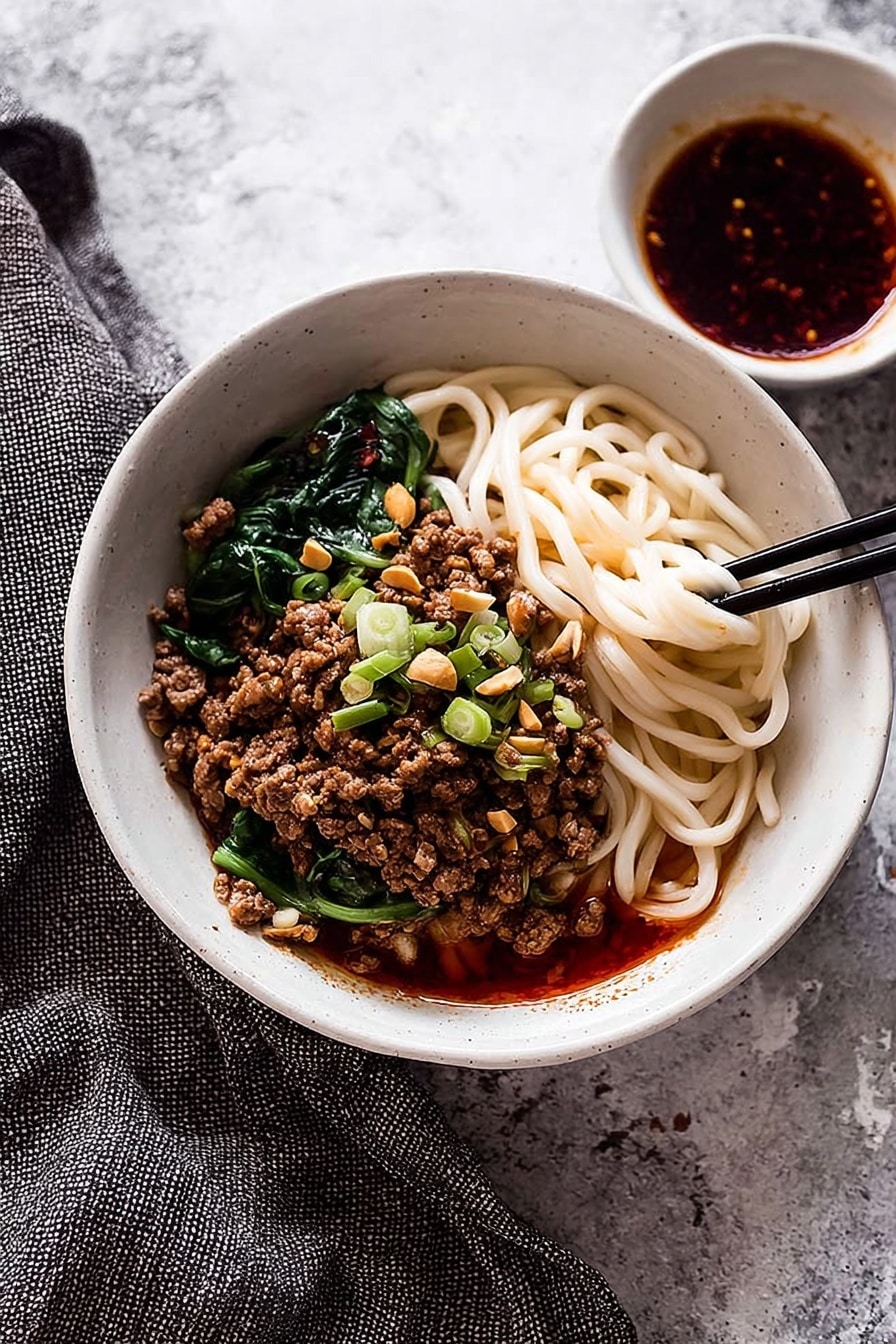 A white bowl filled with three main layers: at the bottom, thick, white noodles partially covered with a red oily sauce; in the middle, a generous layer of browned minced meat topped with chopped green onions and crushed peanuts; and on the top, fresh green leafy vegetables arranged on one side. A pair of wooden chopsticks held by a woman's hand is lifting some noodles, showing their texture and sauce coating. The bowl is placed on a white marbled surface with scattered green onion pieces and crushed peanuts nearby. photo taken with an iphone --ar 2:3 --v 7 - Spicy Dan Dan Noodles with Pork, Dan Dan Noodles, Chinese spicy noodles, Pork noodle stir-fry, Easy Sichuan noodle recipe