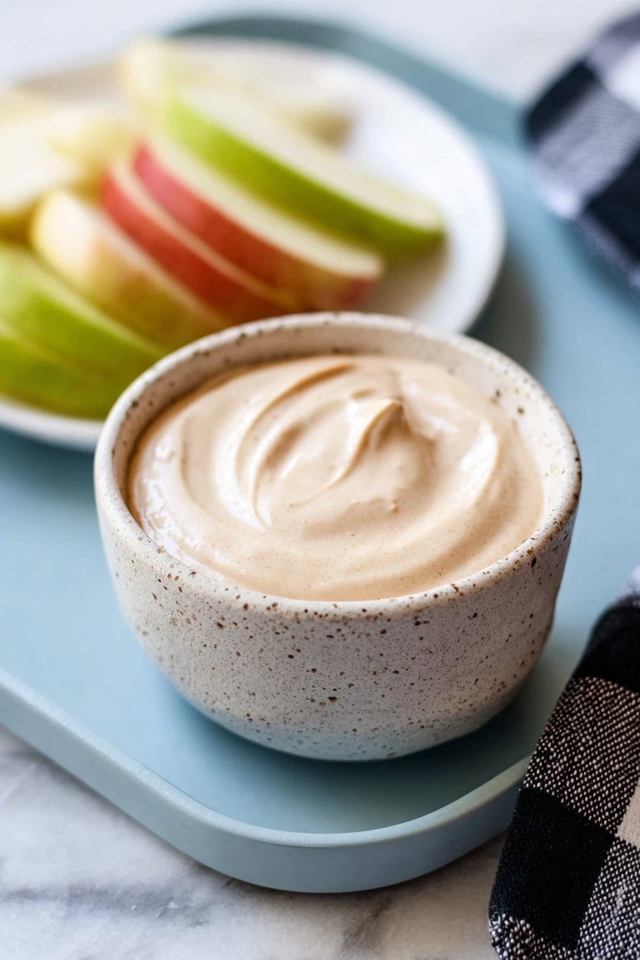 The image shows a small speckled ceramic bowl filled with a smooth, creamy, light beige dip that has soft swirls on the surface. Behind the bowl, there is a white plate with sliced green and red apple wedges, arranged loosely. The bowl and plate are placed on a light blue tray, which rests on a white marbled surface. A black and white checkered cloth is seen partly to the right side. Photo taken with an iphone --ar 2:3 --v 7 - Easy Caramel Apple Dip, caramel apple dip, fall dip recipes, simple apple dip, quick party snacks