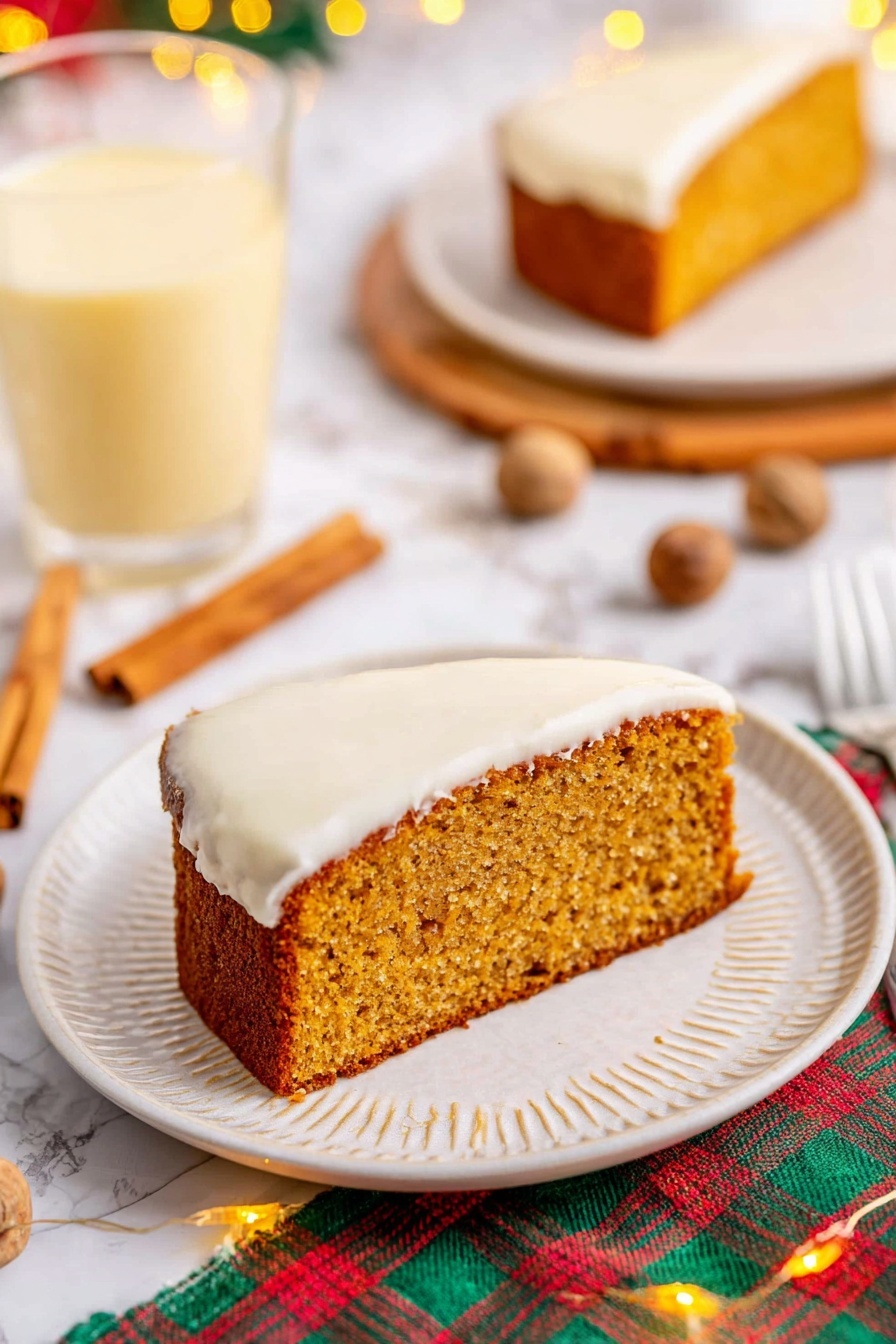 The image shows a single slice of moist, golden-brown cake with a thick layer of smooth white frosting on the bottom side, sitting on a white plate with a subtle textured rim. The cake slice has a slightly rough texture on top, indicating it is soft inside. Around the plate, there are cinnamon sticks and whole nutmegs scattered on a white marbled surface, with a colorful plaid cloth partially visible underneath. In the background, there is a clear glass filled with a light yellow creamy drink, and a second plate holding another slice of the same cake. Small warm fairy lights add a cozy touch to the scene. Photo taken with an iphone --ar 2:3 --v 7 - Eggnog Bread with Spiced Glaze, festive eggnog bread, holiday bread recipes, easy eggnog bread, spiced holiday baked goods