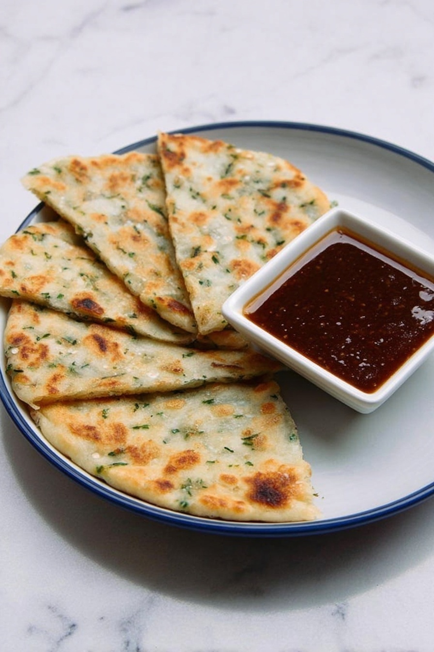 The image shows four pieces of flatbread-like food arranged in a fan shape on a white plate with a blue inside. The flatbreads are light golden-brown with some green herbs visible in the dough, showing a soft and slightly bumpy texture. On the plate, near the upper-right side of the flatbreads, there is a small white square bowl filled with a dark brown sauce that looks smooth and glossy. The plate is placed on a white marbled surface, creating a clean and simple background. photo taken with an iphone --ar 2:3 --v 7 - Chinese Scallion Pancakes, how to make scallion pancakes, crispy scallion pancakes, Chinese street food recipes, homemade scallion pancakes