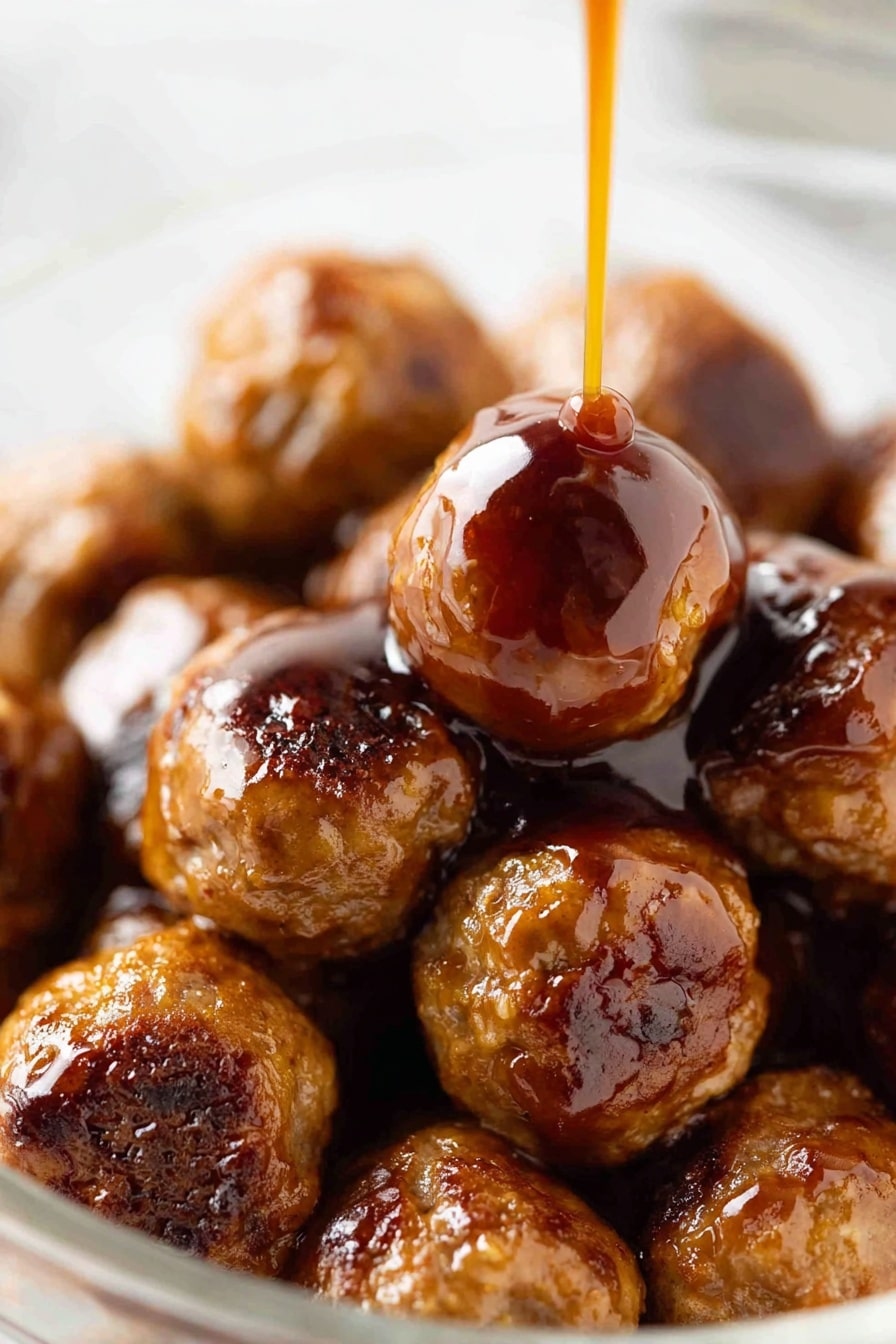 A close-up view of a pile of shiny brown meatballs in a clear bowl on a white marbled surface, with a thick reddish-brown sauce being poured slowly over the top meatball in the center. The meatballs are round and vary slightly in size, with a glazed texture showing small char marks and a glossy finish from the sauce. The sauce is thick and smooth, creating a shiny coating that drips down the sides of the meatballs. The image focuses closely on the meatballs with a soft blurry background. Photo taken with an iphone --ar 2:3 --v 7 - Sweet and Sour Cocktail Meatballs, easy party appetizer, homemade sweet and sour sauce, tasty meatball recipes, finger food for gatherings
