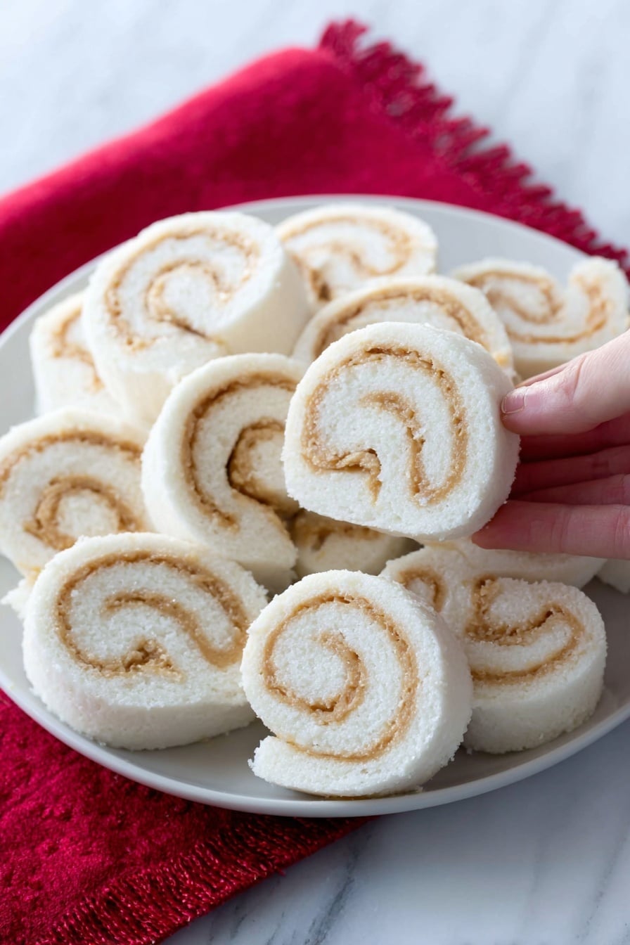 The image shows a white plate filled with soft, spiral-shaped rolls arranged closely together. Each roll has a smooth white outer layer with swirled light tan filling inside, creating a clear contrast. The rolls look fluffy and neatly sliced, with the spirals visible on the sides. The plate is placed on a bright red cloth, and one roll is in the foreground, slightly separated. The background and surface have a white marbled texture. A woman's hand is holding one roll in the top right corner. photo taken with an iphone --ar 2:3 --v 7 - Potato Candy with Peanut Butter Filling, Southern Candy Recipes, Nostalgic Candy Ideas, Easy Candy Recipes, Unique Candy Treats