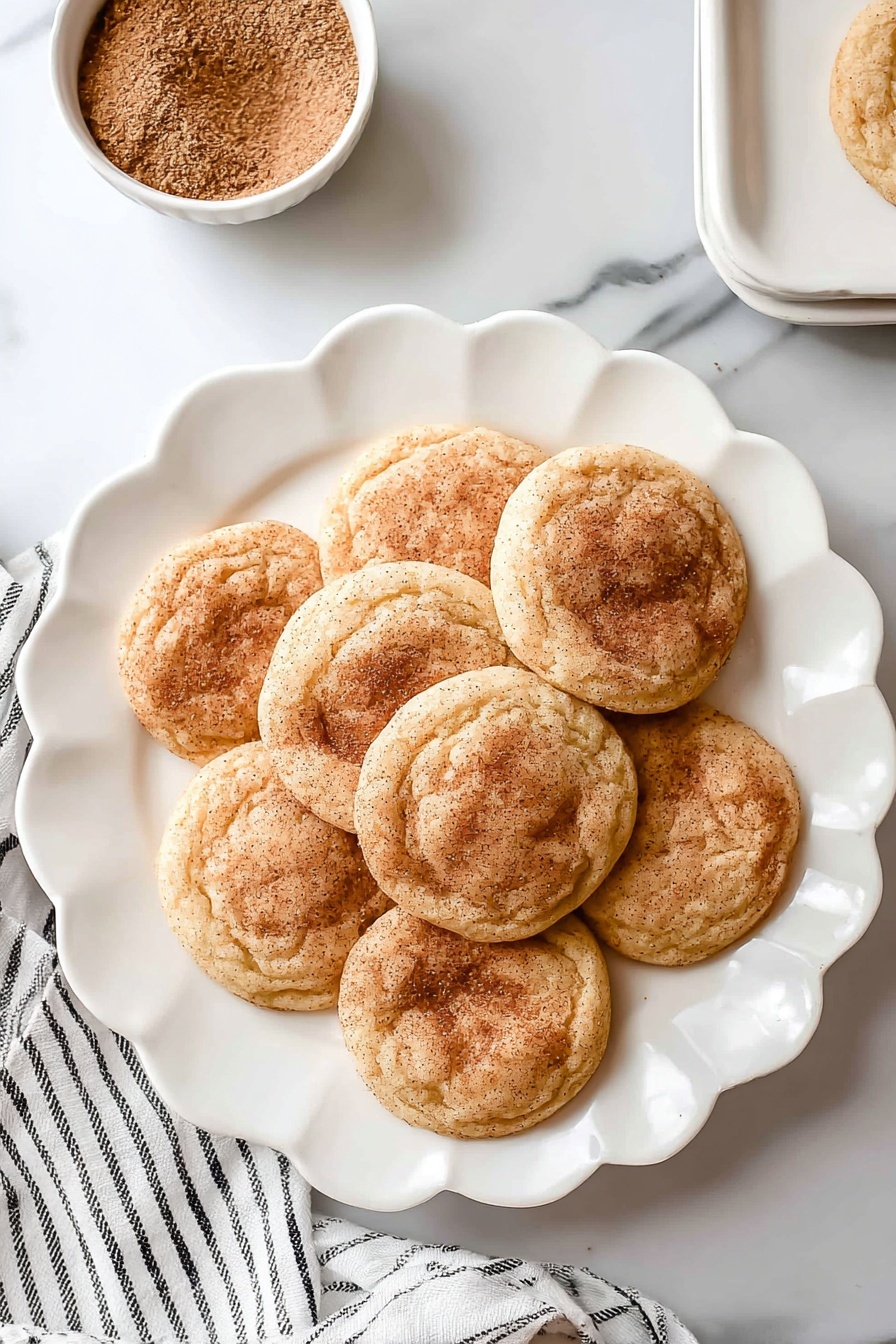 There are seven round snickerdoodle cookies with a light golden brown color and a dusting of cinnamon sugar on top, arranged in a flower shape on a white scalloped plate. The cookies have a soft, slightly wrinkled texture and are placed on a white marbled surface. Near the top left of the plate, there is a small white bowl filled with cinnamon sugar. A white cloth with thin black stripes lays slightly under the plate on the left side. Photo taken with an iphone --ar 2:3 --v 7 - Chewy Snickerdoodle Cookies, cinnamon sugar cookies, soft chewy cookie recipe, homemade snickerdoodle, easy cookie recipes