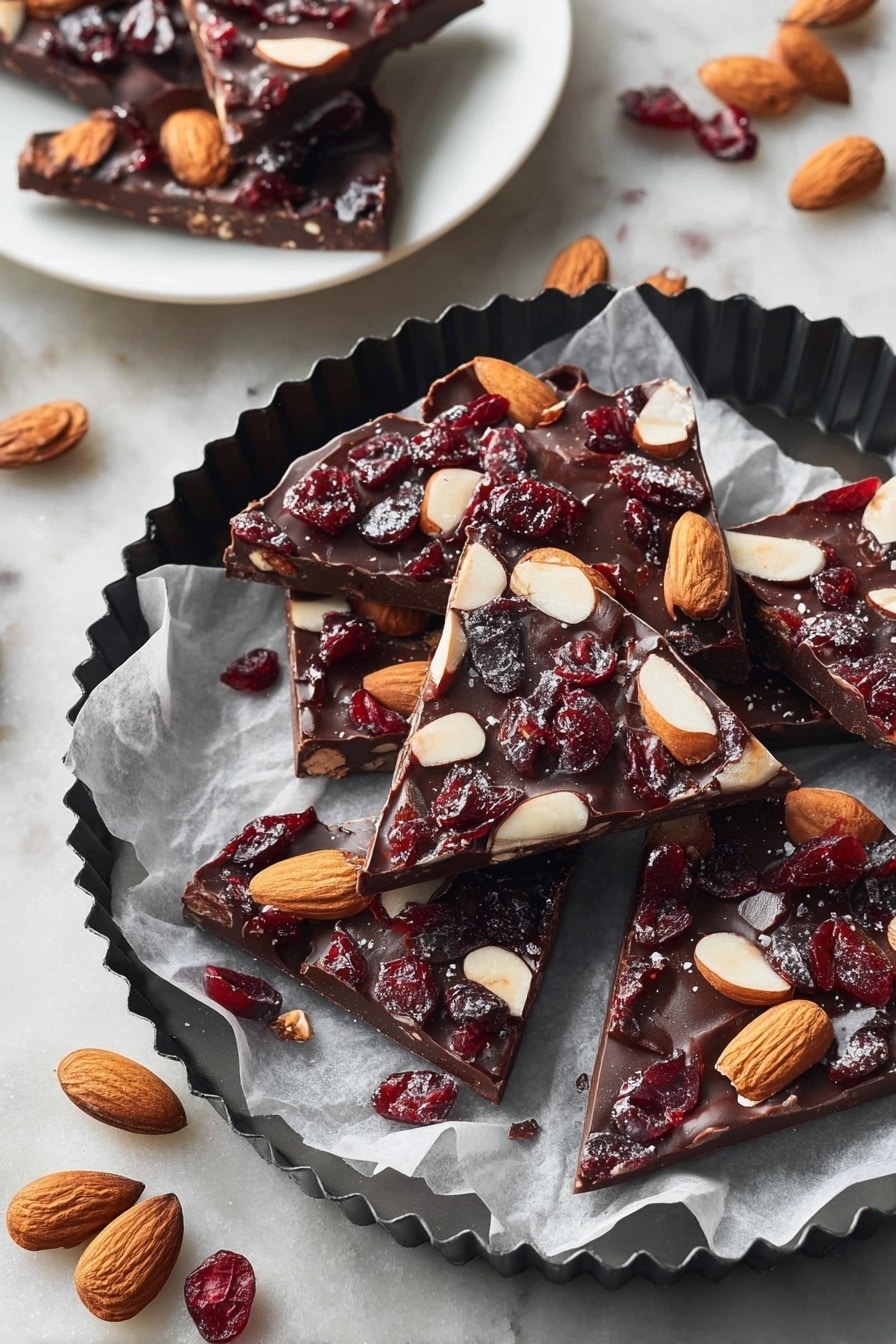 The image shows several pieces of chocolate bark, each with one thick layer of dark brown chocolate topped with whole light brown almonds and dark red dried cranberries scattered evenly. The chocolate pieces are irregular triangles and squares placed on white parchment paper inside a black fluted round pan. Around the pan are whole almonds and dried cranberries on a white marbled textured surface. In the background, a white plate holds three more pieces of chocolate bark, showing the same dark chocolate base with almonds and cranberries. photo taken with an iphone --ar 2:3 --v 7 - Easy Chocolate Bark with Nuts and Fruit, chocolate bark recipe, homemade chocolate toppings, nut and fruit chocolate treat, quick holiday desserts