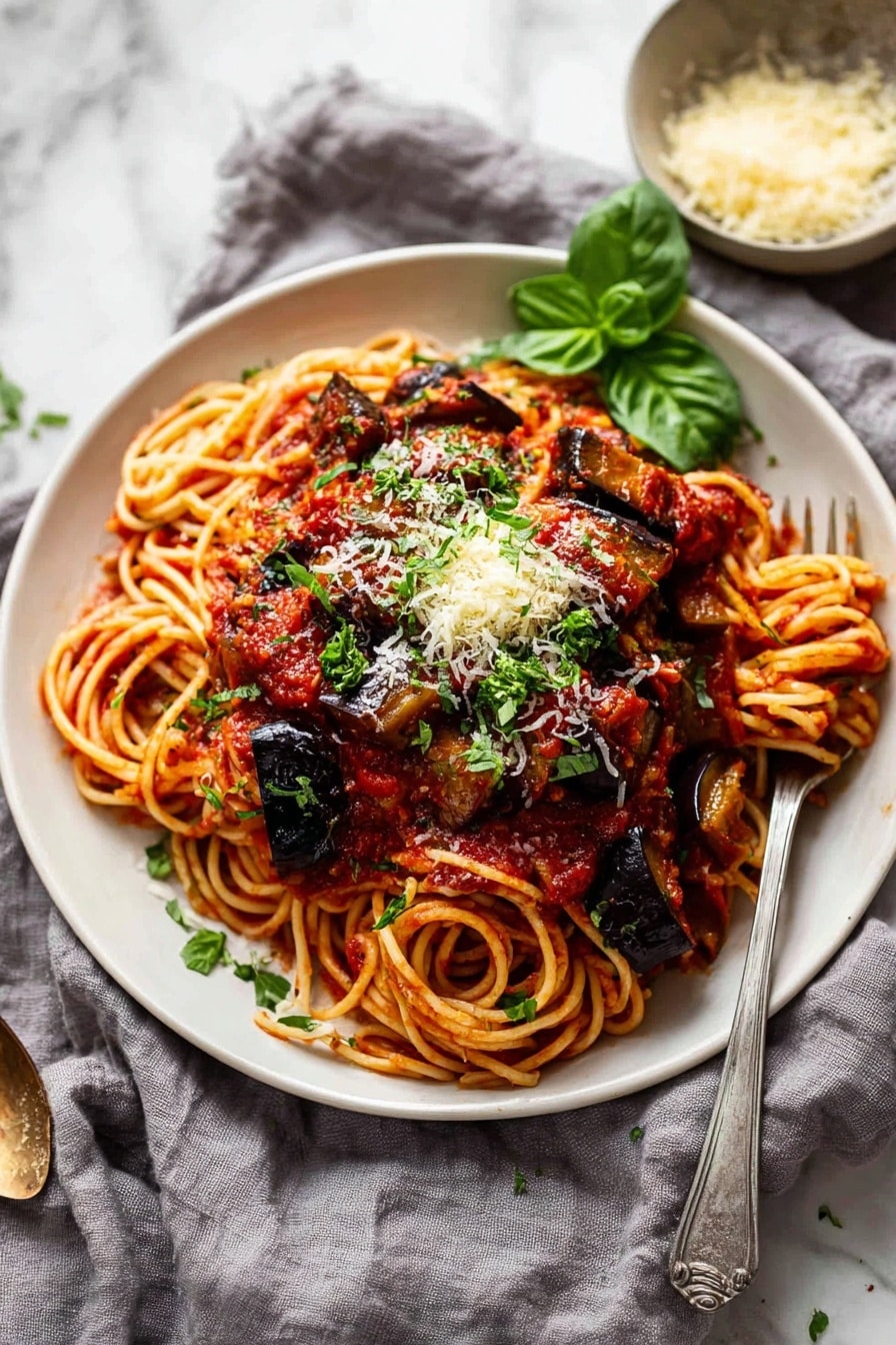 The image shows a close-up of a white bowl filled with spaghetti pasta soaked in rich red tomato sauce, with thick chunks of cooked eggplant scattered on top. The spaghetti strands are tangled at the base and coated evenly with sauce. Large pieces of dark purple eggplant, slightly charred and shiny, sit on the pasta, sprinkled with light yellow grated cheese and small green herb leaves. A silver fork is partly stuck into the pasta, lifting some strands with sauce. The bowl rests on a white marbled surface. Photo taken with an iphone --ar 2:3 --v 7 - Eggplant Pasta alla Norma, Eggplant pasta recipe, Sicilian pasta dishes, Roasted eggplant pasta, Vegetarian pasta recipes