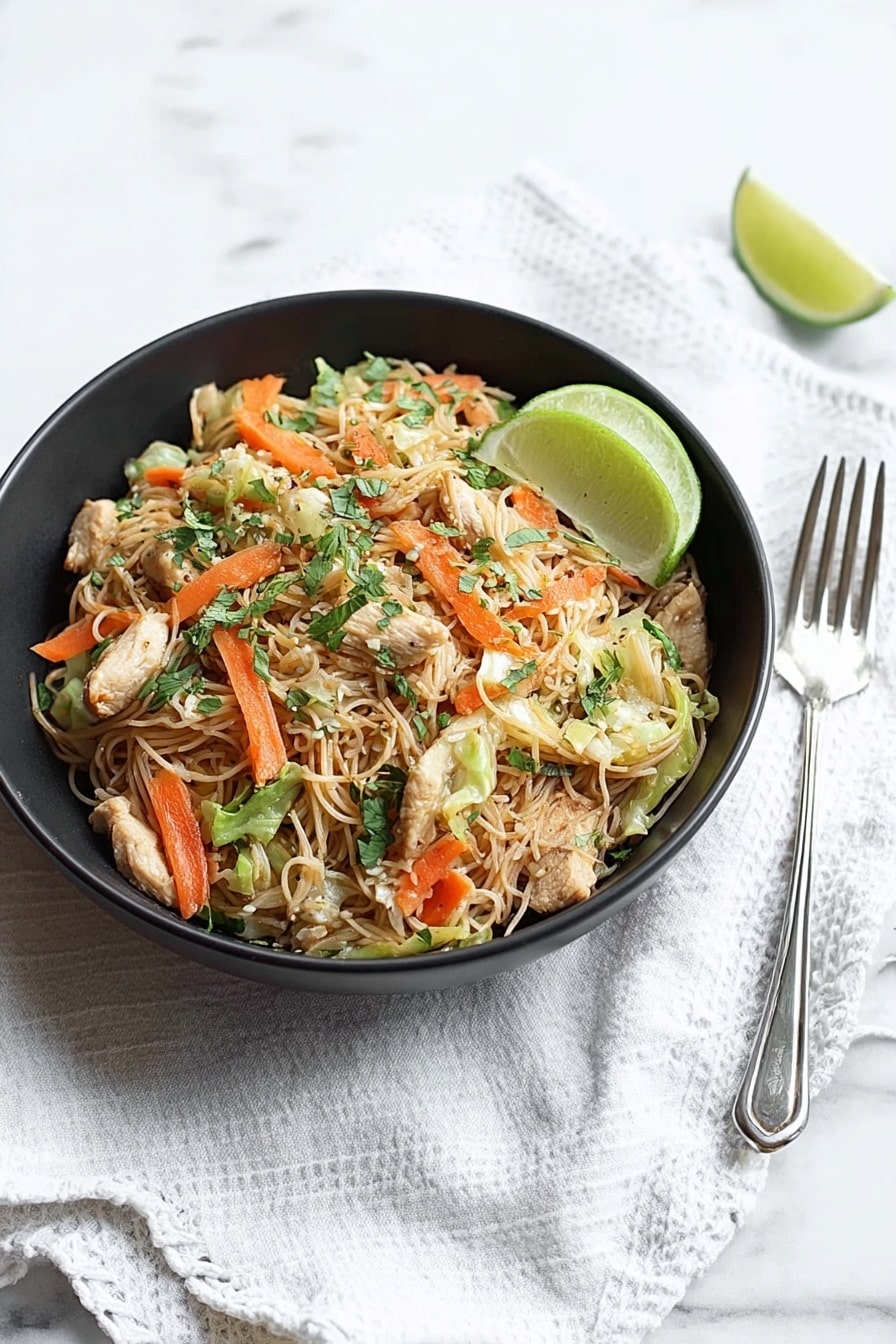 A black bowl sits on a white marbled surface with a white cloth beneath it, filled with thin light brown noodles mixed with small pieces of light beige chicken, bright orange carrot sticks, light green cabbage, and some fresh green herbs scattered on top. A half lime with a pale green inside rests on the right edge of the bowl. To the right of the bowl are a silver spoon and fork placed on the white marbled surface. photo taken with an iphone --ar 2:3 --v 7 - Easy Chicken and Pork Pancit, Filipino noodle dish, quick pancit recipe, savory chicken pork stir-fry, Filipino comfort food