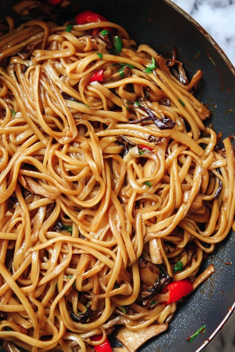 The image shows a close-up of a pan filled with long, shiny noodles coated in a light brown sauce. Mixed among the noodles are small pieces of light tan tofu, dark brown thin mushroom strips, and bright red chili slices, with tiny bits of green herbs scattered throughout. The noodles look soft and glossy, with a slight twist and curl to them, giving a textured, tangled appearance. The pan has a dark surface and the background is changed to a white marbled texture. photo taken with an iphone --ar 2:3 --v 7 - Vegan Kung Pao Pasta, vegan pasta recipes, plant-based Asian pasta, spicy vegan pasta dish, easy vegan dinner ideas