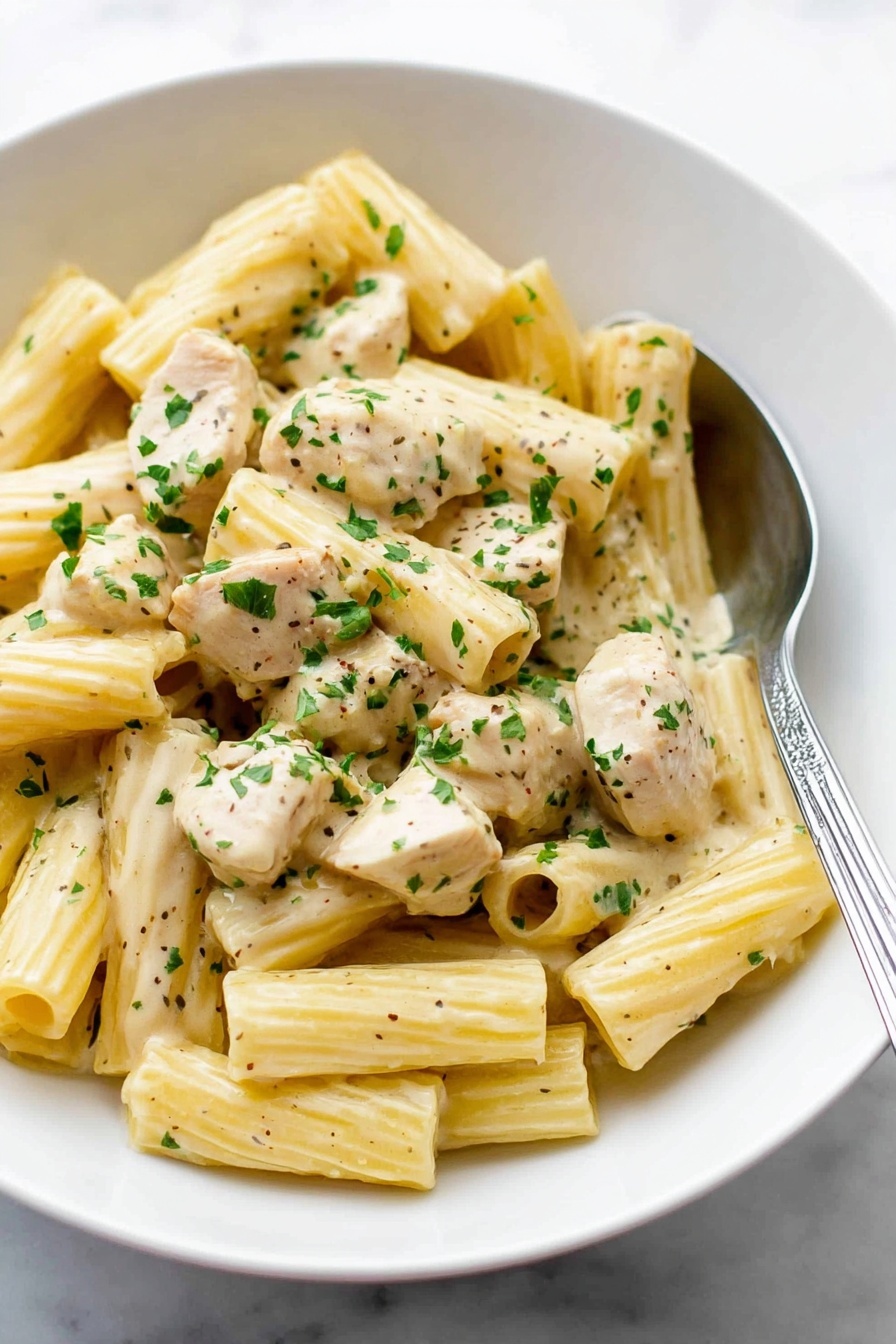 A white bowl filled with creamy rigatoni pasta mixed with chunks of light beige chicken pieces, all coated in a smooth, pale yellow sauce. The pasta is ribbed and tubular, scattered evenly throughout the dish. Small green parsley flakes are sprinkled over the top, adding bright color contrast. A silver spoon rests inside the bowl on the right side, partially under the pasta. The bowl sits on a white marbled surface. Photo taken with an iphone --ar 2:3 --v 7 - Creamy Lemon Chicken Pasta, lemon chicken pasta, creamy pasta with lemon and chicken, quick lemon chicken pasta, easy lemon pasta dinner