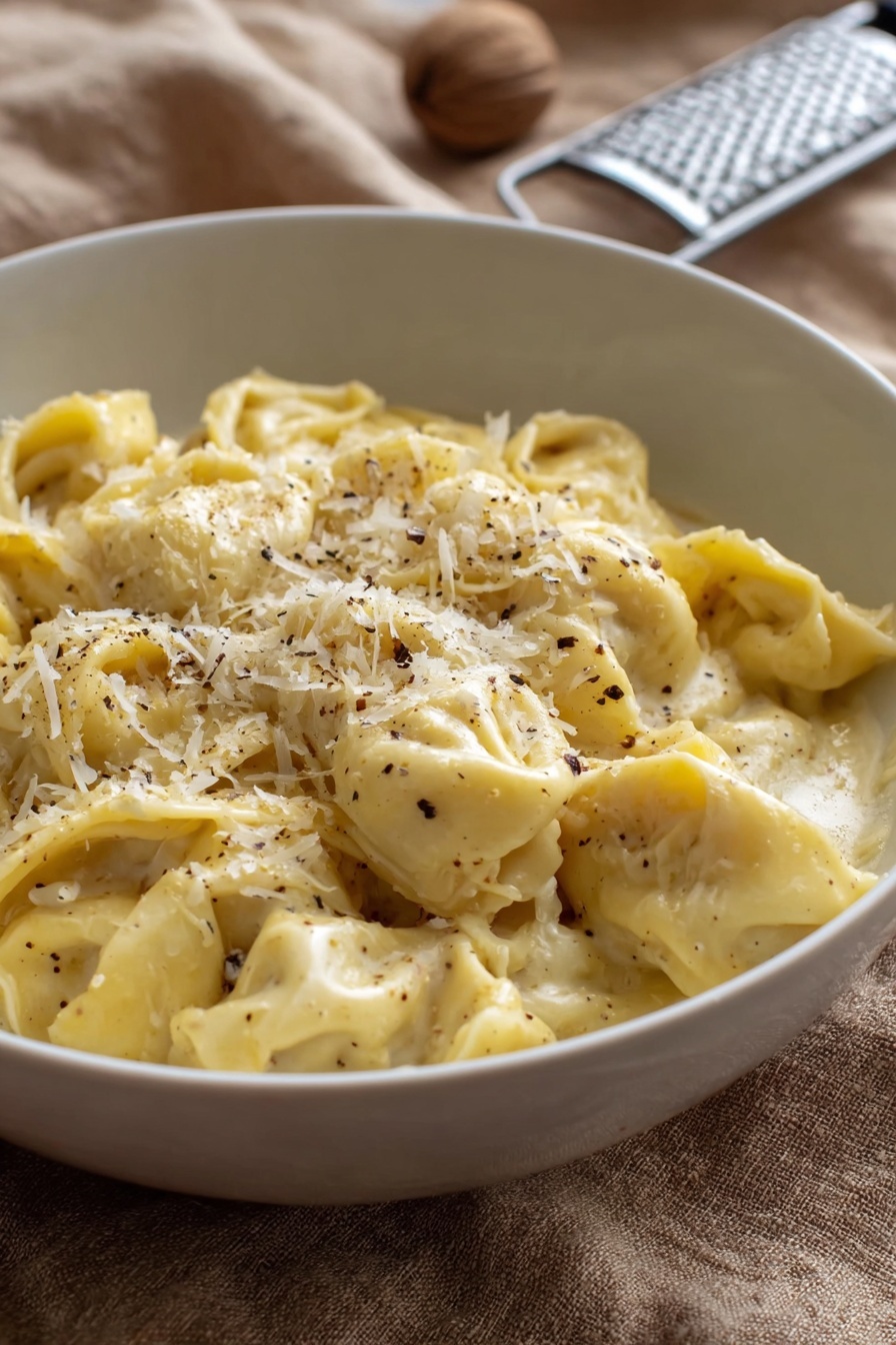 A white bowl filled with about two layers of soft, folded tortellini pasta in a creamy, pale yellow sauce. The pasta is sprinkled with finely grated white cheese and small black pepper flakes, mostly concentrated on the top center. The bowl rests on a tan textured cloth, with a metal grater and a whole nutmeg blurred in the background. The lighting is natural and soft, showing the smooth creamy texture of the sauce and the delicate folds of the tortellini photo taken with an iphone --ar 2:3 --v 7 - Creamy Cheese Tortellini, cheesy tortellini pasta, quick pasta dinner, comfort food recipes, easy cheese tortellini