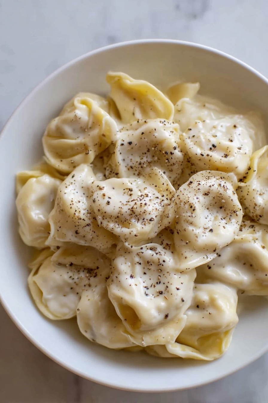A white bowl filled with a single layer of tortellini pasta, covered evenly with a smooth, creamy white sauce that has a slightly shiny texture. The tortellini are pale yellow with soft folds, and black pepper is sprinkled generously on top, adding small dark specks scattered over the sauce. The bowl is placed on a white marbled surface, and the light highlights the soft texture of both the pasta and sauce photo taken with an iphone --ar 2:3 --v 7 - Creamy Cheese Tortellini, cheesy tortellini pasta, quick pasta dinner, comfort food recipes, easy cheese tortellini