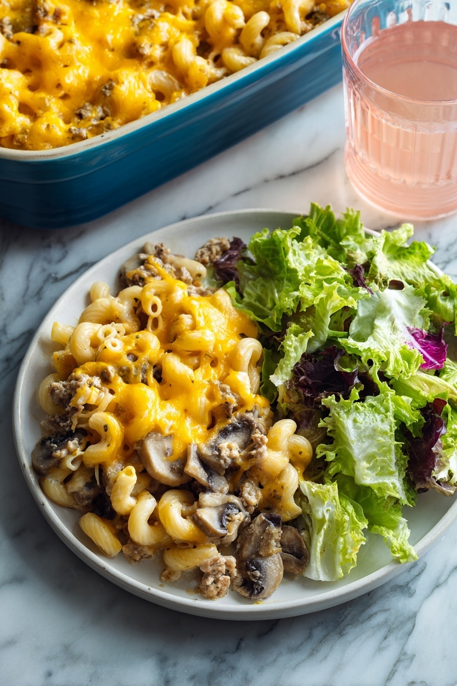 The image shows a white round plate with two main parts: on the left, a layer of baked macaroni mixed with ground meat and sliced mushrooms, all topped with melted bright yellow-orange cheese that looks gooey and soft, and on the right, a fresh green salad made of different lettuce leaves with a mix of light and dark green colors and some purple edges. The plate is set on a white marbled surface, and in the background, there is a blue baking dish filled with more of the macaroni bake and a clear glass of light pink drink. Photo taken with an iphone --ar 2:3 --v 7 - Beef Mushroom Johnny Marzetti Casserole, hearty casserole recipes, comfort food dinners, ground beef mushroom casserole, family-friendly casserole dishes