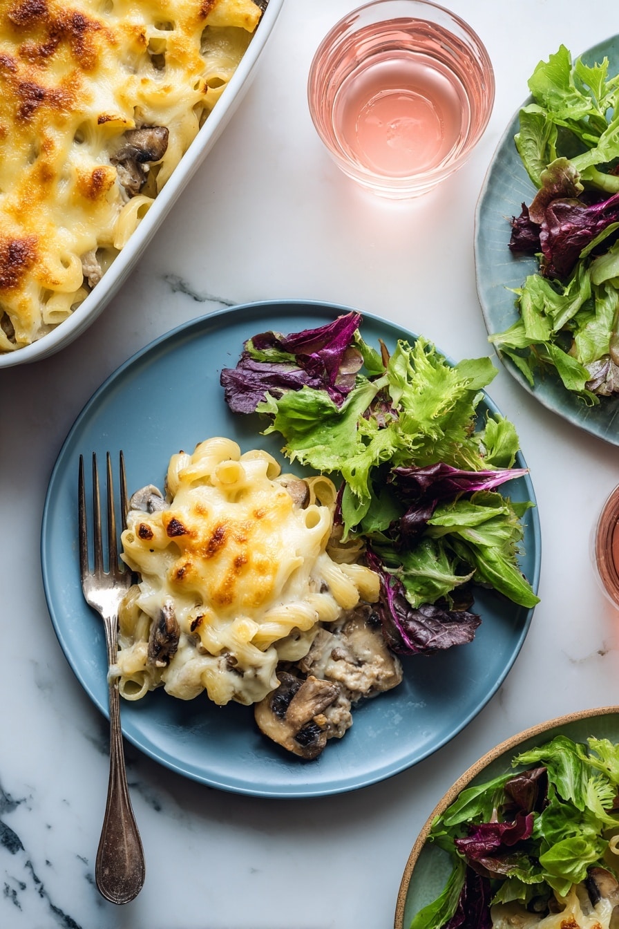 A blue plate holds a serving of baked macaroni and cheese with visible pieces of mushrooms and ground meat under a layer of melted golden-yellow cheese on the left side; on the right side of the plate, there is a fresh green salad made of mixed leafy greens with different shades of green and purple. A silver fork rests on the plate's left edge underneath the macaroni. In the background, a white baking dish filled with more macaroni and cheese sits on a white marbled surface along with a small clear glass of light pink drink. Another plate with leafy greens and another glass of pink drink are partially visible on the right side. photo taken with an iphone --ar 2:3 --v 7 - Beef Mushroom Johnny Marzetti Casserole, hearty casserole recipes, comfort food dinners, ground beef mushroom casserole, family-friendly casserole dishes