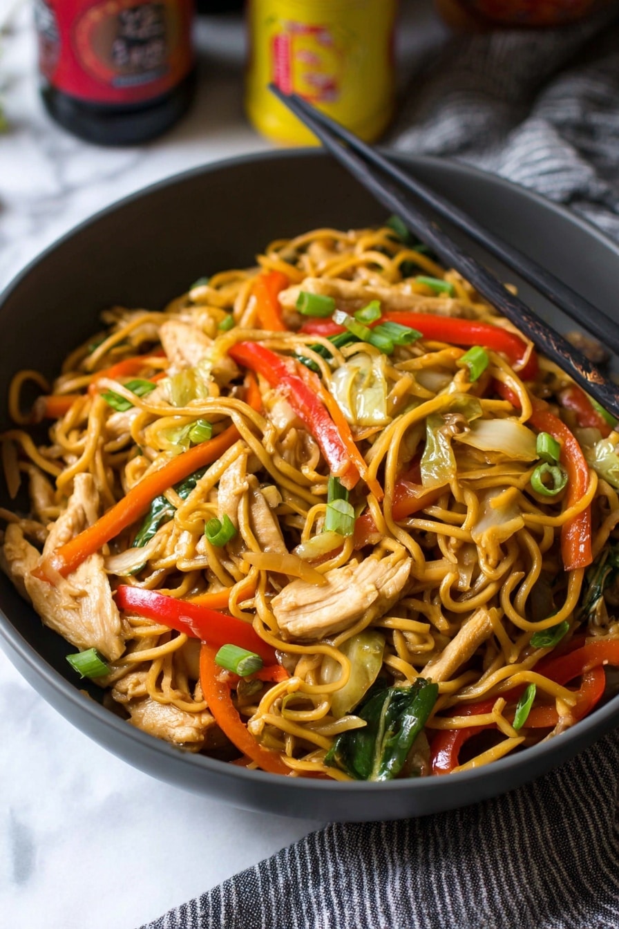 A close-up view of a dark gray bowl filled with stir-fried noodles mixed with thin strips of light beige chicken pieces, bright red bell pepper slices, green scallion pieces, thin orange carrot strips, light green cabbage, and sliced beige mushrooms. Black chopsticks rest on the right edge of the bowl, while the bowl sits on a white marbled surface with part of a gray striped cloth to the right. The background includes some blurred bottles with yellow and red labels. photo taken with an iphone --ar 2:3 --v 7 - Chicken Yakisoba Stir-Fry, Chicken Yakisoba, Japanese stir-fry recipes, easy chicken stir-fry, quick Asian noodle dishes