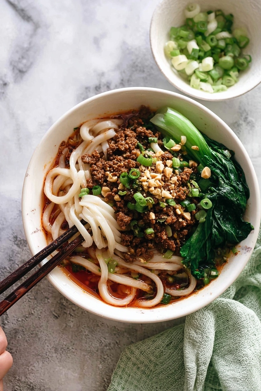The image shows a white bowl filled with three main layers: at the bottom, thick white noodles soaked in a reddish-brown sauce, in the middle, crumbled cooked meat with chopped green onions and crushed peanuts sprinkled on top, and on the side, bright green leafy vegetables placed neatly. A pair of dark wooden chopsticks is picking up some noodles from the bowl. Next to the bowl, there is a small white bowl with sliced green onions, and a light green towel is folded in the background. The whole scene is set on a white marbled surface. photo taken with an iphone --ar 2:3 --v 7 - Spicy Dan Dan Noodles with Pork, Dan Dan Noodles, Chinese spicy noodles, Pork noodle stir-fry, Easy Sichuan noodle recipe