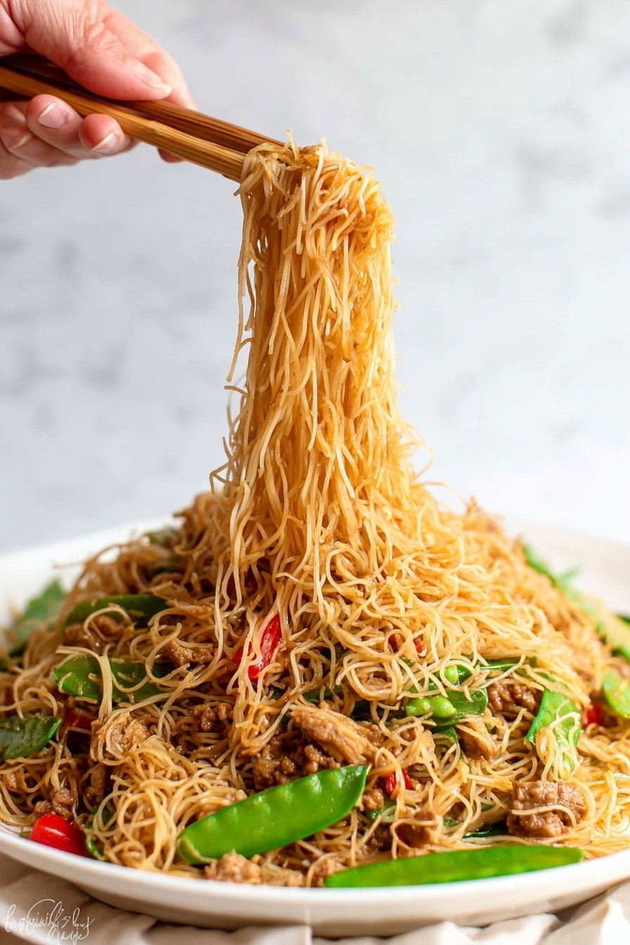 The image shows a white plate full of thin noodles mixed with small pieces of cooked meat, green snow peas, and red bell pepper slices. The noodles are light brown and soft, forming the top and main layer, while the vegetables and meat are scattered throughout, adding pops of green and red colors. A woman's hand is holding wooden chopsticks lifting a portion of the noodles above the plate, with the noodles hanging down in thin strands. The background has a white marbled texture. photo taken with an iphone --ar 2:3 --v 7 - Singapore Curry Noodles, easy Singapore curry noodles, Asian noodle recipes, quick curry noodle dish, flavorful noodle recipes