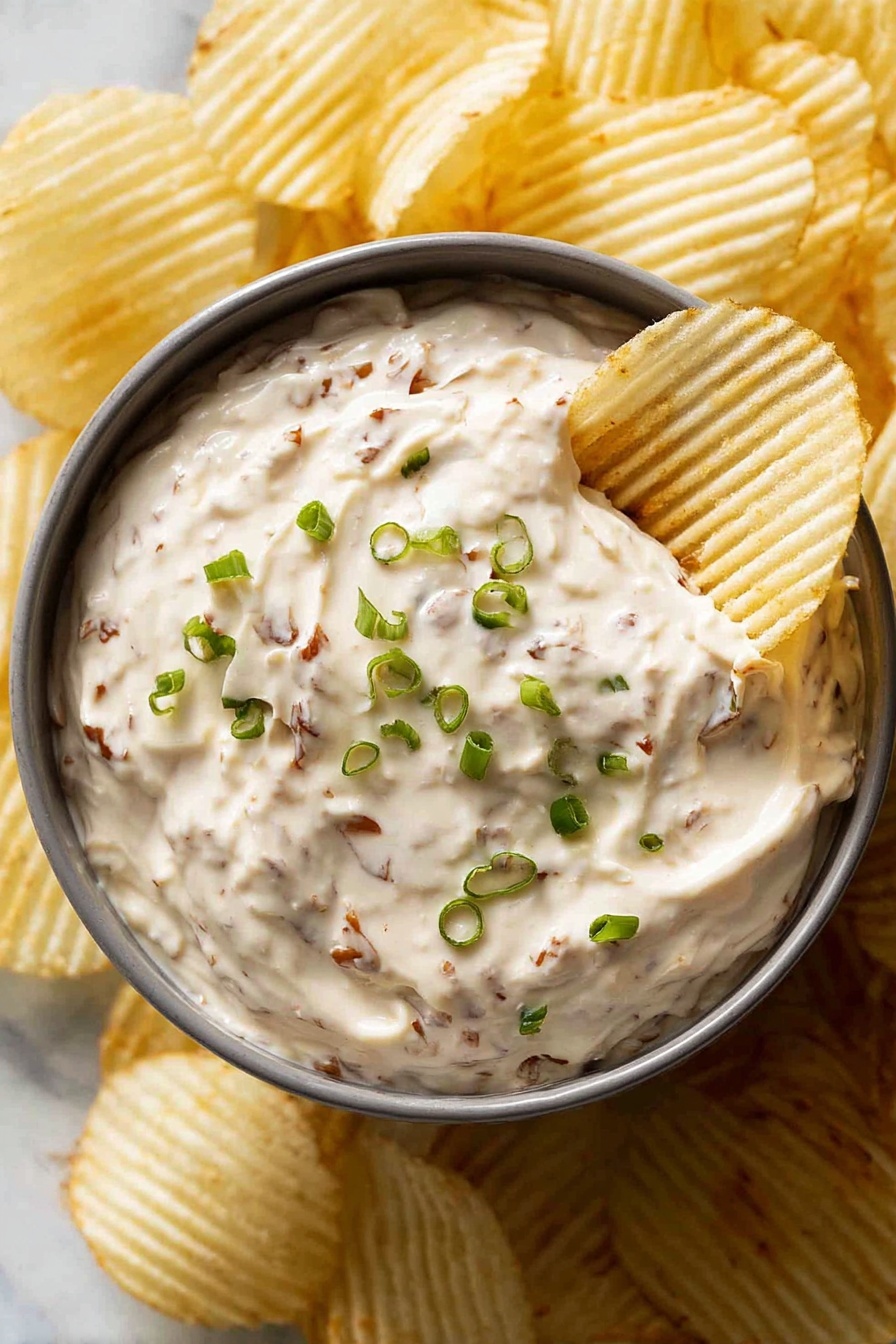A gray round bowl filled with creamy dip that has visible small brown bits throughout, topped with finely chopped green onions scattered on the surface. A white ridged potato chip is dipped into the creamy mixture on the right side of the bowl. The bowl is surrounded by more white ridged potato chips, all arranged on a white marbled surface. photo taken with an iphone --ar 2:3 --v 7 - French Onion Dip, homemade French onion dip, easy French onion dip, creamy onion dip, savory onion dip