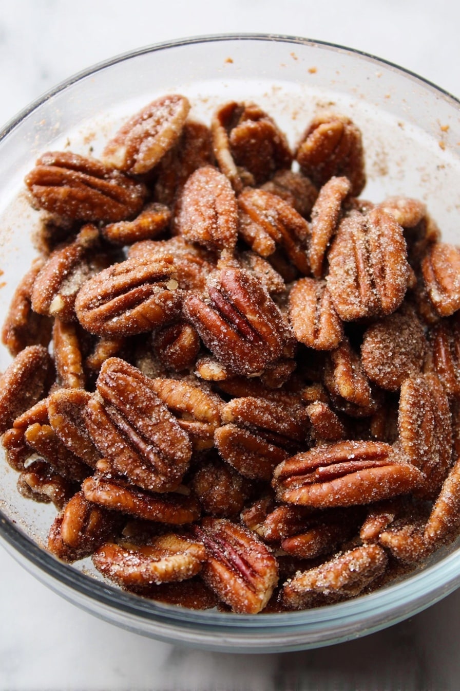 A clear glass bowl is full of many toasted pecan halves, each nut showing a rough, crinkled surface with a shiny brown color covered lightly with a layer of fine sugar crystals and cinnamon dust. The bowl sits on a white marbled surface. photo taken with an iphone --ar 2:3 --v 7 - Cinnamon Candied Pecans, candied pecans recipe, easy pecan snack, sweet spicy nuts, holiday pecans