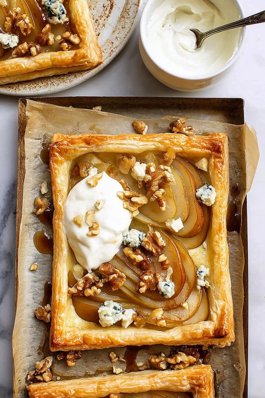 The image shows a golden brown puff pastry tart with a raised edge surrounding soft, thinly sliced caramelized pears arranged in an overlapping pattern. On top of the pears, there are small chunks of blue cheese and scattered walnut pieces adding texture and color contrast. A dollop of creamy white yogurt or cream is placed on one corner of the tart. The tart sits on parchment paper which lines a baking tray with some walnut pieces and cheese crumbs around it. Nearby, a small white ceramic bowl with a spoon inside contains more white cream. The background is a white marbled surface. photo taken with an iphone --ar 2:3 --v 7 - Caramelized Pear and Blue Cheese Tart, pear and blue cheese tart, easy savory tart, caramelized pear appetizer, blue cheese puff pastry