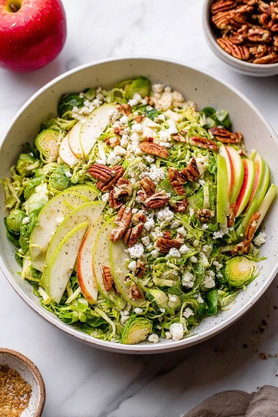 A large white bowl filled with a fresh salad featuring several layers: the base layer is thinly sliced green Brussels sprouts, topped with light green and white strips of apple. Scattered on top are golden-brown pecans and small white crumbles of cheese. The ingredients have a fresh, slightly rough texture with visible black pepper sprinkled over. The bowl sits on a white marbled surface, and in the background, a red apple and a small bowl of pecans are visible. Photo taken with an iphone --ar 2:3 --v 7 - Fall Brussels Sprout Salad with Apple and Blue Cheese, healthy fall salads, easy Brussels sprouts salad recipe, seasonal apple and blue cheese salad, quick autumn side dish