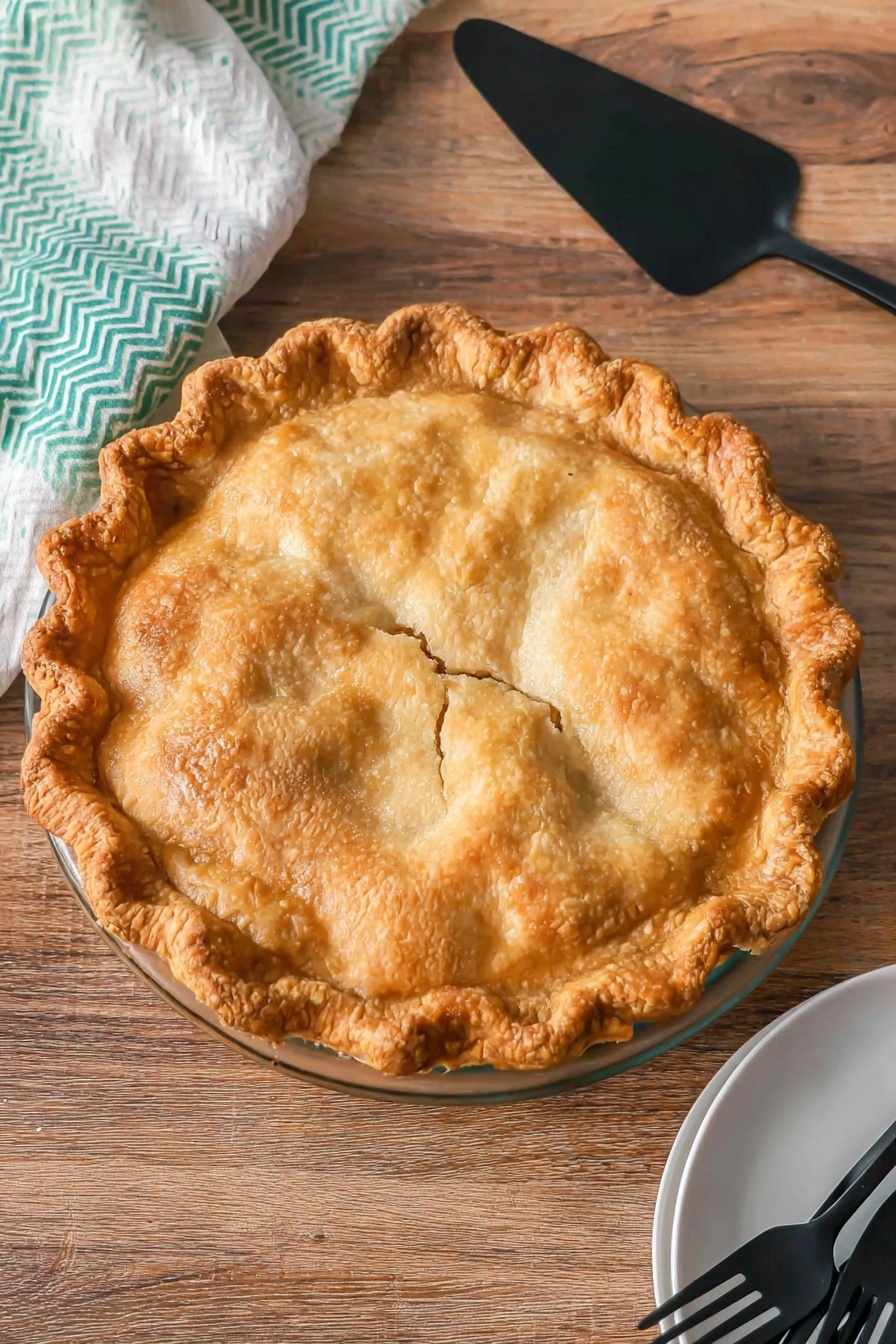 A whole golden-brown pie with a flaky, bubbly crust and a crimped edge sits centered on a clear glass pie dish. The crust has a slightly uneven, homemade texture with a few small cracks near the middle, showing a hint of the filling below. The pie is placed on a wooden surface, above which rests a black pie server with a matte finish. In the bottom right corner, there is a white plate holding three black forks, and to the left, a white cloth with a green chevron pattern peeks into the frame. photo taken with an iphone --ar 2:3 --v 7 - Easy Pear Pie, Pear Pie, Quick Pear Dessert, Easy Fruit Pie, Simple Pear Tart