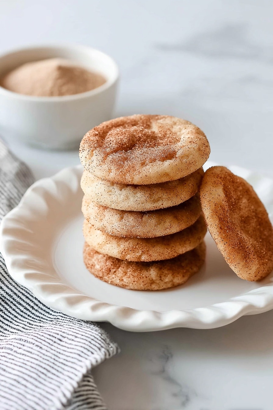 A stack of eight round, light brown cookies with a dusting of cinnamon sugar on top sits in the center of a white plate with scalloped edges. One cookie lies flat behind the stack, showing its darker cinnamon spots. The white plate is placed on a white marbled surface with a folded white and black striped cloth partially visible on the left. In the top left corner, a small white bowl filled with a light brown powder is slightly out of focus. photo taken with an iphone --ar 2:3 --v 7 - Chewy Snickerdoodle Cookies, cinnamon sugar cookies, soft chewy cookie recipe, homemade snickerdoodle, easy cookie recipes