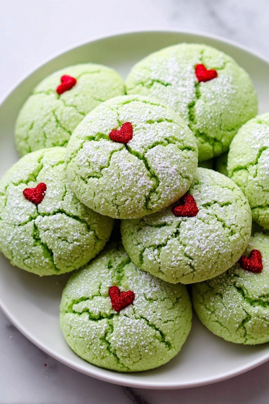 The image shows a white plate filled with round green cookies that have a cracked texture on top. Each cookie has a light dusting of white powder and a tiny red heart decoration placed slightly off-center on the top surface. The cookies are stacked closely together on the plate, which sits on a white marbled background. Photo taken with an iphone --ar 2:3 --v 7 - Grinch Cookies with Peppermint and Green Food Coloring, festive holiday cookies, mint-flavored Christmas cookies, easy green holiday treat, peppermint cookie recipe