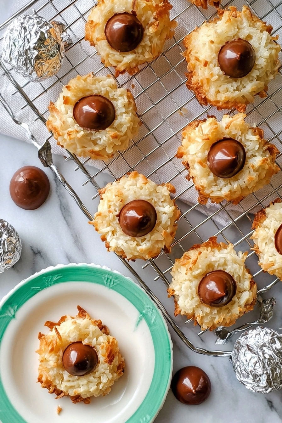 The image shows seven round coconut cookies with a light golden-brown crispy outside and white, shredded coconut texture in the middle layer. Each cookie has a single smooth milk chocolate drop placed in the center on top. The cookies are arranged on a silver cooling rack that rests on a white rectangular marble slab. Around the rack, there are several extra chocolate drops scattered on the white marbled surface. A woman's hand is holding one cookie on a white plate with a green rim, and a few silver-wrapped chocolate candies are placed nearby. The overall setting is bright with a white marbled background. photo taken with an iphone --ar 2:3 --v 7 - Coconut Macaroon Blossoms with Chocolate Kisses, coconut macaroon cookies, chocolate kiss cookies, festive coconut treats, easy holiday desserts