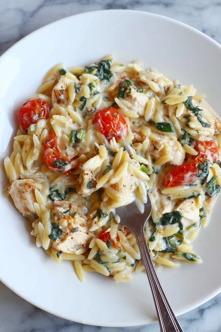 A white round plate holds a creamy pasta dish composed of orzo pasta mixed with small chunks of light brown cooked chicken pieces and bright red cherry tomatoes. There are dark green leafy bits, possibly spinach or herbs, spread evenly throughout the dish. The sauce looks smooth and creamy, coating the pasta and ingredients in a soft white layer. A silver fork is placed inside the pasta near the bottom center of the plate. The plate sits on a white marbled surface photo taken with an iphone --ar 2:3 --v 7 - Creamy Chicken Orzo Bake, easy chicken orzo casserole, cheesy chicken pasta bake, hearty chicken and orzo dish, baked chicken with tomatoes and spinach