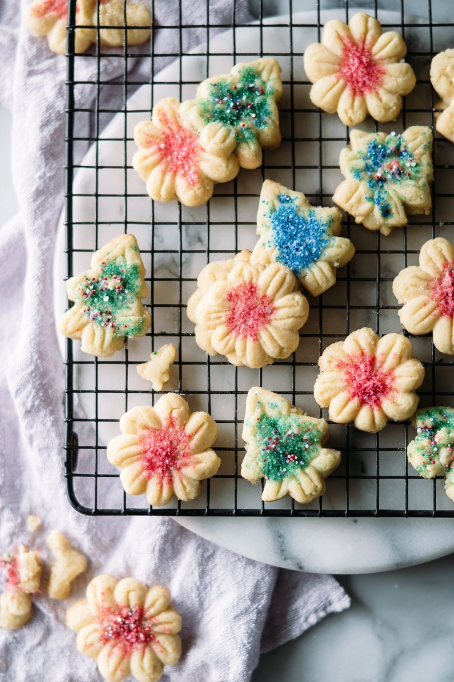 The image shows small, shortbread cookies in two shapes: flower-like rounds and Christmas trees, arranged on a round white marble board and a black wire cooling rack placed on a white marbled surface with a white cloth underneath. The cookies have a light beige color and are decorated with colored sugar sprinkles—red on the round flower-shaped cookies and blue or green on the Christmas tree-shaped cookies. The cookies appear slightly textured with small ridges from the mold, and they are scattered loosely on both the marble board and the wire rack, creating a casual but festive look. Photo taken with an iphone --ar 2:3 --v 7 - Butter Spritz Cookies, buttery spritz cookies, classic spritz cookies, easy spritz cookie recipe, almond spritz cookies