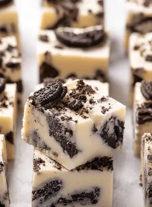 The image shows a close-up of thick square pieces of cookies and cream fudge layered in white creamy base mixed with broken chocolate cookie bits. Each piece has a smooth, pale off-white layer filled with dark brown cookie chunks inside and baked bits scattered on top, creating a textured look with irregular shapes. The fudge pieces are placed closely together on a white marbled surface, highlighting the contrast between the creamy white fudge and dark cookies. Photo taken with an iphone --ar 2:3 --v 7 - White Chocolate Oreo Fudge, easy Oreo fudge recipe, no-bake Oreo dessert, quick white chocolate fudge, simple Oreo fudge