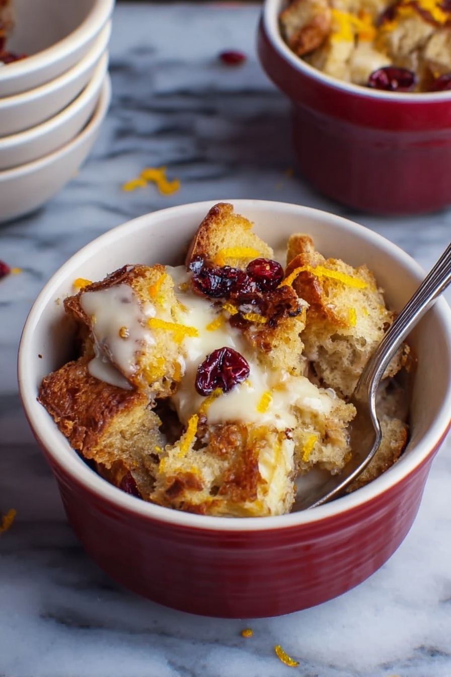A small white bowl filled with several layers of toasted bread chunks that are golden brown and crispy on the edges. The bread pieces are mixed with creamy, slightly melted white sauce or cream that sits on top and around the bread. There are dark red dried berries scattered on top and some thin orange zest strips add a pop of color. A silver spoon is inserted inside the bowl on the right side. The bowl is placed on a white marbled surface with a small stack of empty white bowls in the background. photo taken with an iphone --ar 2:3 --v 7 - Cranberry Easy Bread Pudding with Toffee, cranberry bread pudding, festive bread pudding, holiday dessert recipes, easy bread pudding with cranberries
