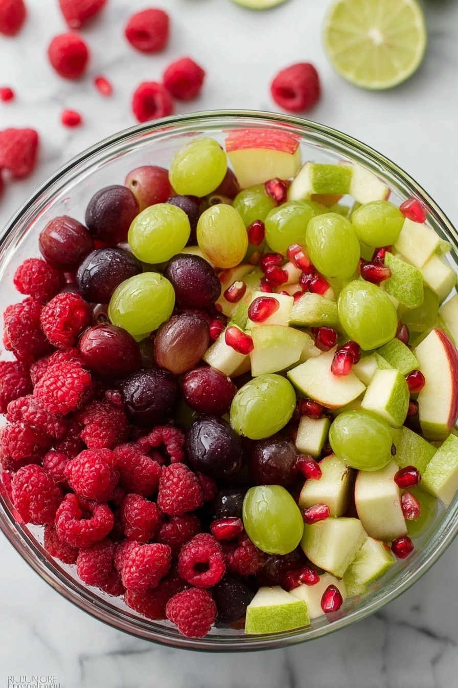 A clear glass bowl filled with a colorful fruit salad sits on a white marbled surface. The salad has multiple layers made up of red raspberries, dark purple grapes, light green grapes, small pieces of green kiwi with black seeds, light yellow apple slices with a red skin edge, and shiny red pomegranate seeds scattered on top. The fruits create a mix of vibrant reds, purples, greens, and yellows with smooth and textured surfaces. Photo taken with an iphone --ar 2:3 --v 7 - Festive Christmas Fruit Salad, holiday fruit salad, Christmas fruit salad, easy holiday desserts, healthy Christmas treat