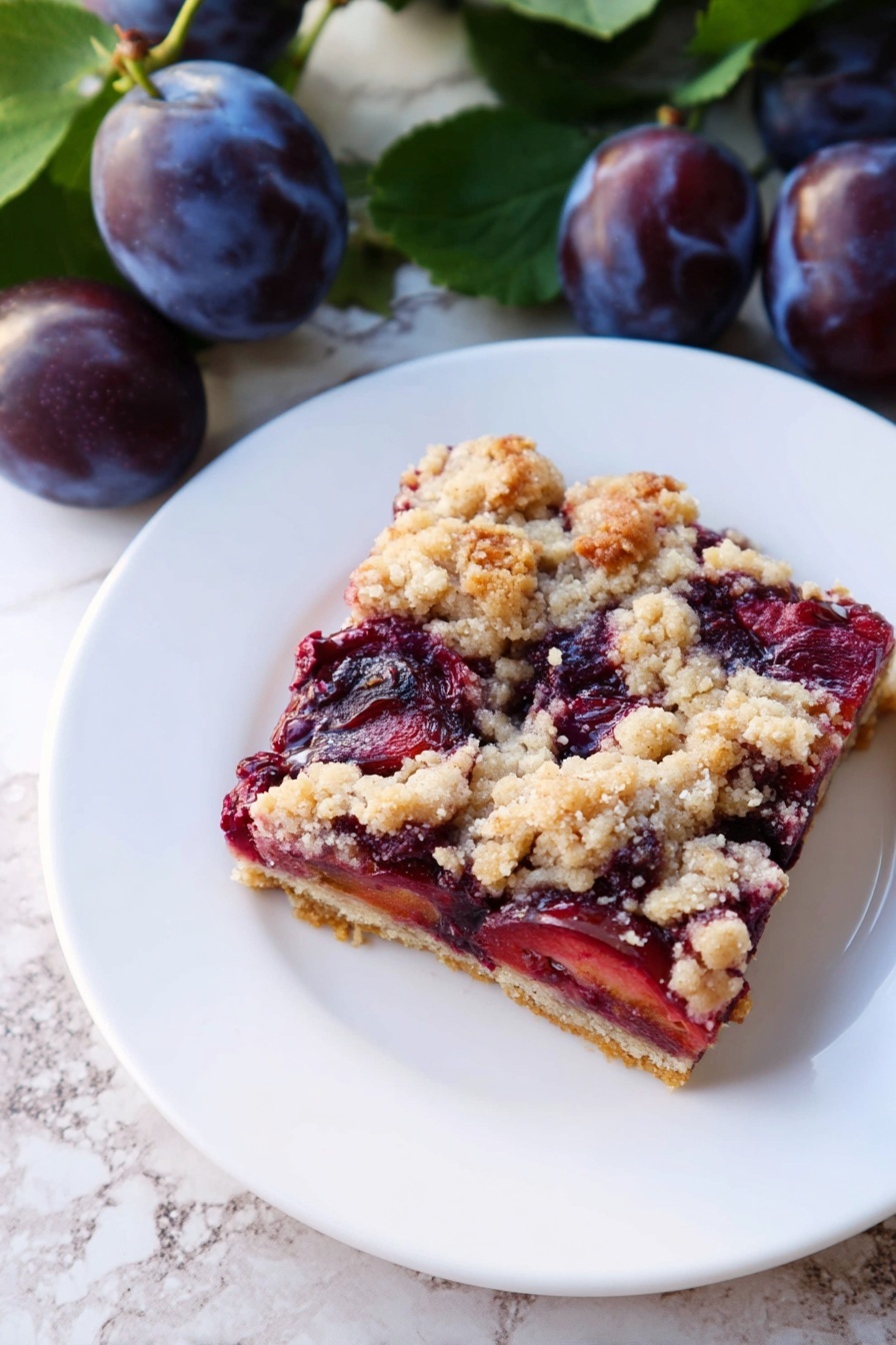 A square slice of a fruit crumble bar sits on a white plate. The bottom layer is a light golden crust, topped with a thick layer of dark red and purple cooked plums showing a soft texture. The top layer is a crumbly streusel with uneven pieces of pale beige and light brown. The plate is placed on a white marbled surface, next to fresh whole dark purple plums with green leaves. Photo taken with an iphone --ar 2:3 --v 7 - German Plum Cake with Yeast Crust, Zwetschgenkuchen, Traditional German Plum Dessert, Yeast Crust Plum Cake, German Fruit Pastry