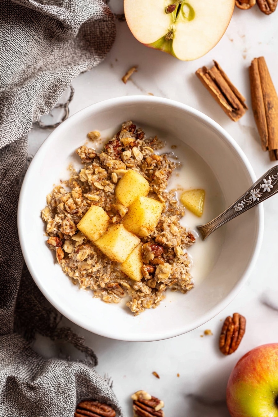 A white bowl on a white marbled surface holds a serving of baked oatmeal with visible oats and pieces of cooked apple mixed throughout. The oatmeal is light brown with a crumbly texture and small bits of nuts adding roughness. On top, there are a few chunks of golden yellow apple pieces. A silver spoon with a floral handle rests inside the bowl, partly submerged in white milk around the oatmeal. Nearby on the surface are whole apples, cinnamon sticks, and pecans, with a gray woven cloth folded beside the bowl. photo taken with an iphone --ar 2:3 --v 7 - Cinnamon Apple Baked Oatmeal, healthy baked oatmeal with apples, cozy breakfast ideas, easy make-ahead breakfast recipes, nourishing oatmeal breakfast