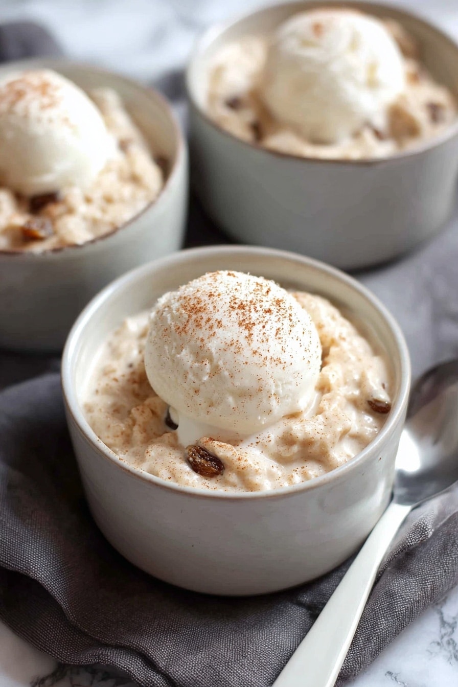 A white bowl filled with a thick creamy oatmeal mixture that has a light tan color with soft, chunky texture and dark brown raisins mixed throughout. On top of the oatmeal is a single scoop of smooth, pale white ice cream with a light dusting of brown cinnamon powder. A silver spoon is partially submerged in the oatmeal on the right side of the bowl. The bowl sits on a soft pink cloth with a white marbled surface underneath. Photo taken with an iphone --ar 2:3 --v 7 - Creamy Cinnamon Rice Pudding, rice pudding with cinnamon, easy rice pudding recipe, cozy dessert with raisins, baked rice pudding