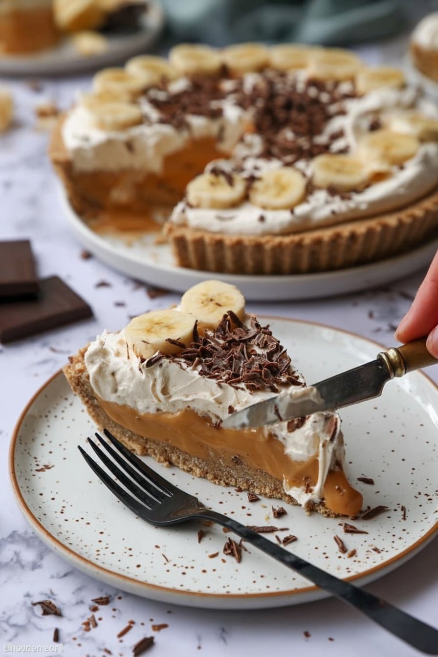 A slice of pie sits on a white plate with brown speckles, showing three layers: a crunchy light brown crust, a smooth caramel-colored filling with banana slices, and a creamy white topping sprinkled with dark chocolate curls. Behind it, the rest of the pie with the same layers is on a white plate, with one slice missing. A woman’s hand holds a silver knife with some pie filling and cream on it. A black fork rests on the plate near the slice. The background is a white marbled surface with some chocolate shavings scattered around. Photo taken with an iphone --ar 2:3 --v 7 - No-Bake Banoffee Pie, easy banoffee dessert, no-bake pie recipe, caramel banana pie, quick dessert ideas