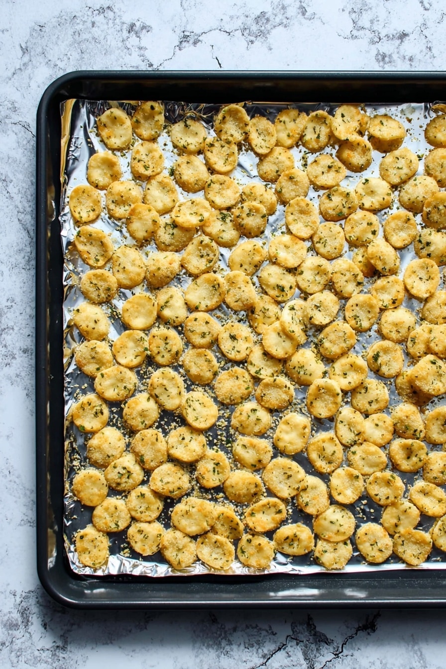 A black baking tray lined with shiny aluminum foil holds a single flat layer of small, round, golden-brown crackers sprinkled with green herbs. The crackers are spread evenly, with slight overlaps in places, showing a crispy and slightly rough texture. The background features a white marbled surface that contrasts with the baking tray. photo taken with an iphone --ar 2:3 --v 7 - Ranch Oyster Crackers, Easy snack recipes, Party snack ideas, No-bake cracker snacks, Savory snack recipes