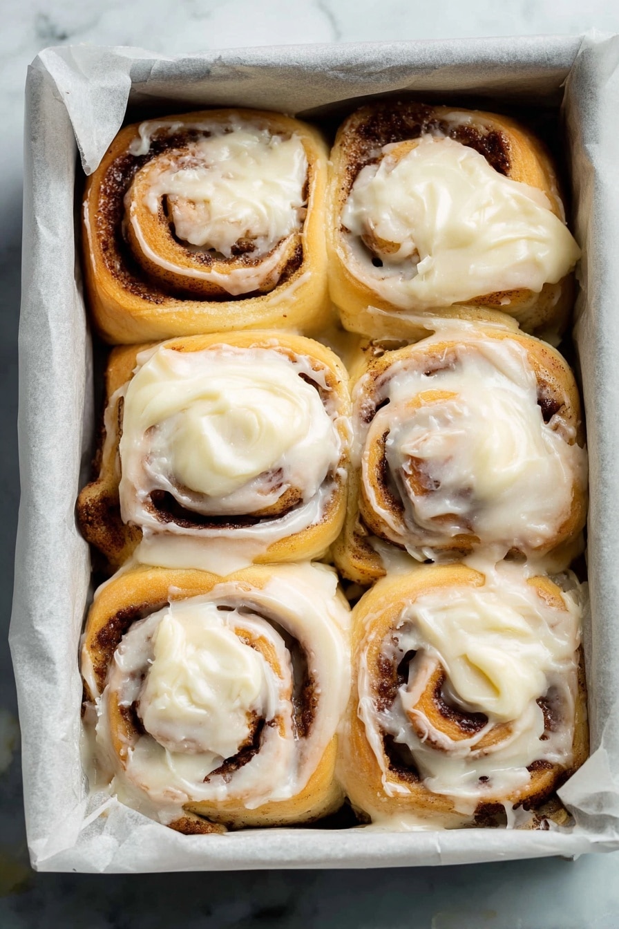 This image shows eight cinnamon rolls in a square baking pan lined with white parchment paper, placed on a white marbled surface. Each roll has two visible layers: the base layer is soft, light golden dough spiraled with a dark brown cinnamon filling, and the top layer is thick, creamy white frosting spread unevenly, melting slightly into the rolls. The rolls are packed closely together, creating a warm, inviting look with some frosting softly dripping between them. Photo taken with an iphone --ar 2:3 --v 7 - Ultimate Fluffy Cinnamon Rolls, easy cinnamon roll recipe, homemade cinnamon rolls, soft cinnamon rolls with cream cheese frosting, best cinnamon rolls