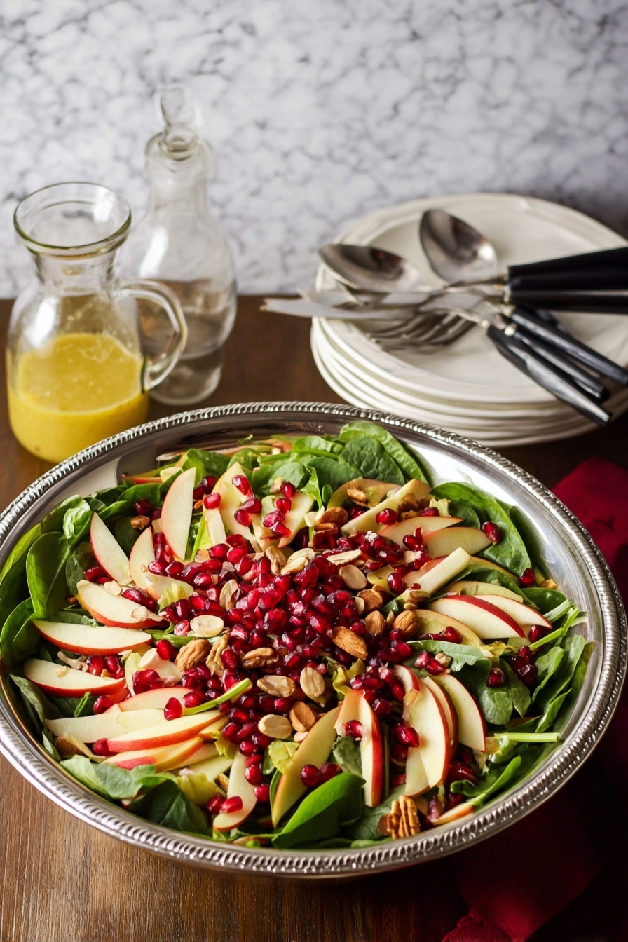 A large silver bowl holds a fresh salad with three main layers: the bottom layer is dark green spinach leaves with smooth texture, the middle layer is thin, light cream and red slices of apple arranged evenly over the spinach, and the top layer has bright red pomegranate seeds and small pieces of light tan nuts sprinkled throughout. The bowl is on a wooden surface with a white marbled texture background. Nearby are two silver serving spoons, a small glass jar of yellow dressing, and a stack of white plates with black-handled forks. photo taken with an iphone --ar 2:3 --v 7 - Apple Cranberry Spinach Salad, healthy fruit and vegetable salad, easy holiday salad recipe, fresh winter salad, nutritious spinach salad