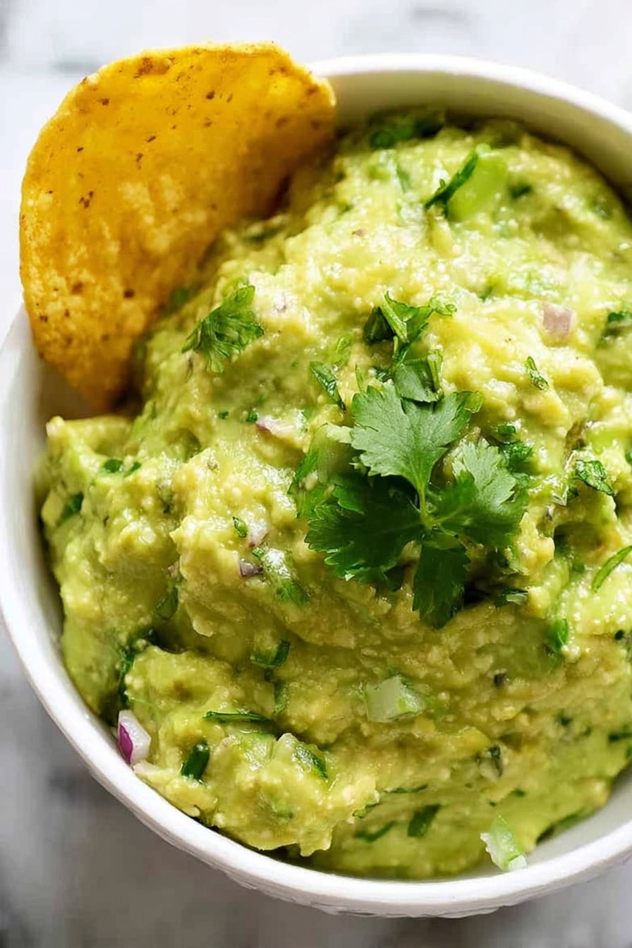 A close-up view of a white bowl filled with creamy, chunky pale green guacamole mixed with small bits of green herbs and red onion, topped with fresh bright green cilantro leaves. On the left side of the bowl, a golden yellow corn chip is partially dipped into the guacamole. The bowl sits on a white marbled surface. photo taken with an iphone --ar 2:3 --v 7 - Easy Homemade Guacamole, quick guacamole recipe, simple guacamole dip, fresh mexican guacamole, how to make guacamole