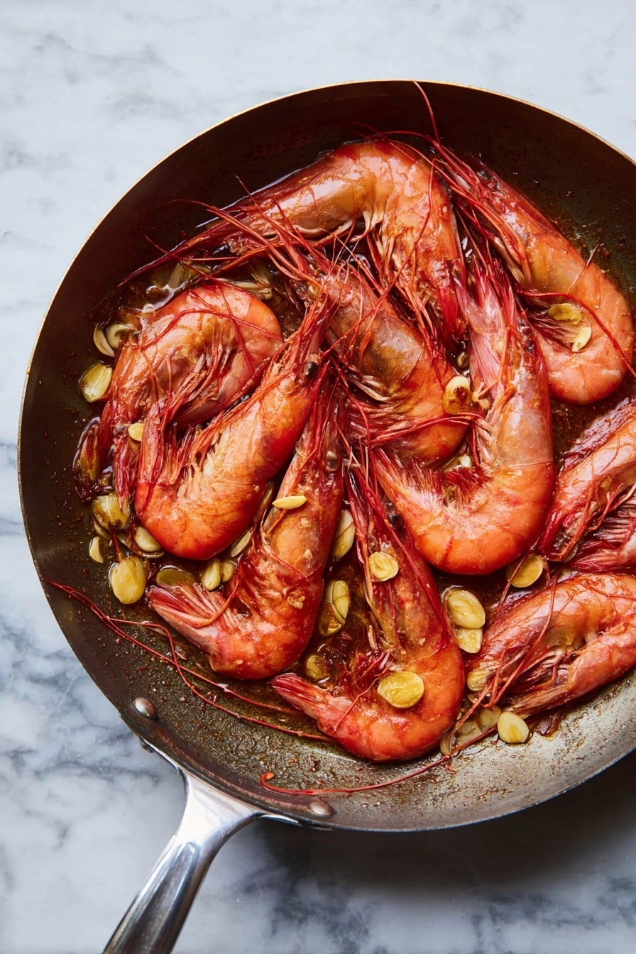 A frying pan filled with cooked whole red prawns layered over scattered golden brown slices of sautéed garlic. The prawns are arranged in a slightly overlapping way, showing their curved, shell-covered bodies, long antennae, and black eyes. The pan has a shiny metal handle extending to the side, and the background is a white marbled surface. photo taken with an iphone --ar 2:3 --v 7 - Spanish Garlic Shrimp, garlic shrimp recipe, quick seafood dishes, Spanish appetizers, easy shrimp recipes