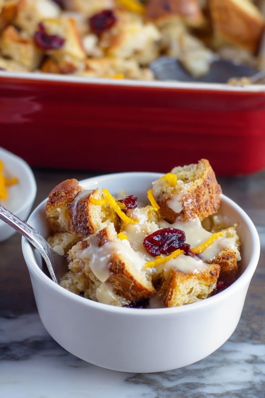 A small white bowl filled with several uneven layers of toasted bread pieces that are golden brown and crispy, mixed with a creamy, light-colored sauce spread unevenly throughout, topped with a few dried red cranberries and thin strips of bright orange zest, placed on a white marbled surface with a silver spoon inside the bowl, a larger baking dish filled with similar bread pudding in the background, photo taken with an iphone --ar 2:3 --v 7 - Cranberry Easy Bread Pudding with Toffee, cranberry bread pudding, festive bread pudding, holiday dessert recipes, easy bread pudding with cranberries