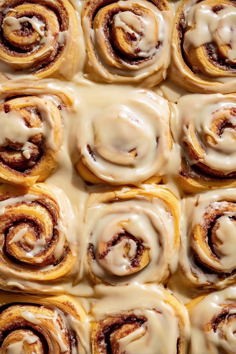 The image shows a close-up view of twelve cinnamon rolls tightly placed in a white pan, each roll having a spiral shape with layers of soft, golden-brown dough and dark cinnamon filling in between. The rolls are generously covered with smooth, creamy, light beige icing that spreads over the top and drips down slightly onto the sides and edges of each roll. The texture of the dough looks soft and fluffy, while the cinnamon filling adds a rich contrast in color. The light brown icing has a glossy finish, adding a sense of sweetness and warmth to the whole scene. The white marbled surface beneath complements the warm tones of the cinnamon rolls. photo taken with an iphone --ar 2:3 --v 7 - Gingerbread Cinnamon Rolls with Cream Cheese Frosting, festive cinnamon rolls, holiday breakfast recipes, gingerbread desserts, holiday cinnamon roll recipe