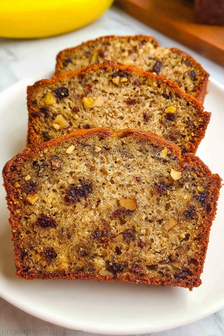Three slices of moist banana nut bread with a golden brown crust are arranged in a row on a white plate. The bread has a dense, textured inside, showing chopped nuts and dark raisins evenly spread throughout each slice. The background is a white marbled surface with a yellow banana blurred in the back. photo taken with an iphone --ar 2:3 --v 7 - Moist Banana Bread with Walnuts and Raisins, banana bread with walnuts and raisins, fluffy banana bread recipe, homemade banana bread, easy banana bread with nuts and dried fruit