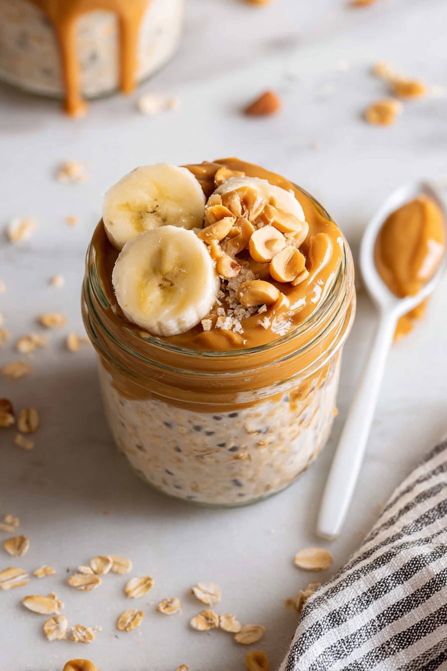 The image shows a clear glass jar filled with three layers: a bottom layer of creamy oatmeal with visible oats and seeds, a middle layer of smooth peanut butter spread thickly, and a top layer decorated with three half-moon slices of banana and sprinkled with chopped peanuts. The jar is placed on a white marbled surface with scattered oats and banana slices around. A white spoon lies beside the jar, and a striped cloth is partially visible on the right side. photo taken with an iphone --ar 2:3 --v 7 - Healthy Protein Overnight Oats, healthy breakfast ideas, high-protein oats, quick and easy breakfast recipes, nutritious overnight oats