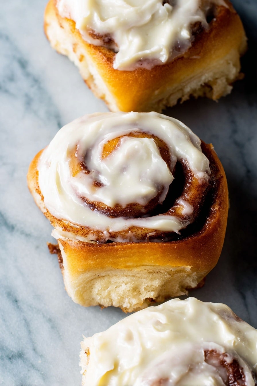 The image shows three cinnamon rolls with soft golden-brown spiral layers of dough swirled with cinnamon filling. Each roll is topped with a creamy white frosting that looks thick and smooth, spread unevenly with some parts thicker than others. The cinnamon rolls are placed on a white marbled surface that contrasts with their warm colors. The focus is close up, making the texture of the dough and frosting very clear. Photo taken with an iphone --ar 2:3 --v 7 - Ultimate Fluffy Cinnamon Rolls, easy cinnamon roll recipe, homemade cinnamon rolls, soft cinnamon rolls with cream cheese frosting, best cinnamon rolls