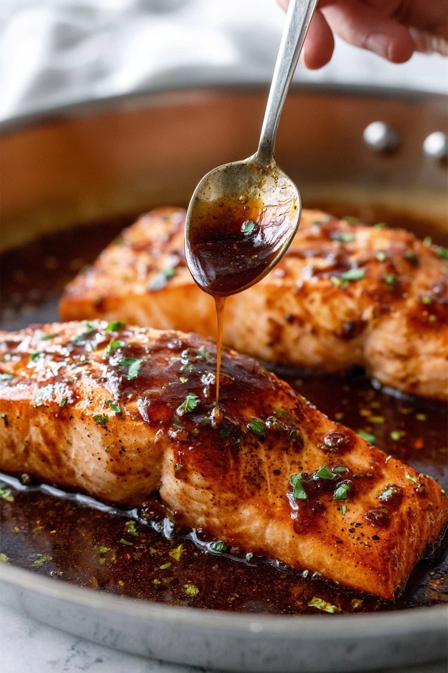 Two thick salmon fillets with a shiny dark brown sauce cover. The top layer is glossy, sticky sauce with small green herb bits sprinkled over. The salmon’s surface is textured with light char marks and a golden brown color under the sauce. A woman's hand holds a metal spoon above the front fillet, drizzling more sauce from the spoon onto the fish. The fillets sit in a shallow pool of the dark sauce within a metal pan. The background has a soft, white marbled texture. photo taken with an iphone --ar 2:3 --v 7 - Honey Garlic Salmon, honey garlic salmon, quick salmon recipe, healthy salmon dinner, easy seafood recipes
