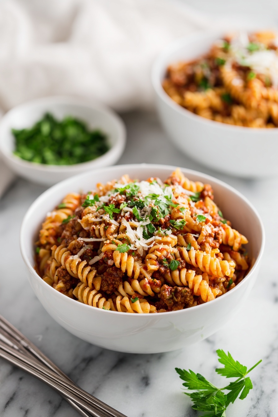 A white bowl filled with a layered pasta dish made of ruffled-edge pasta mixed with a red tomato sauce, ground meat, and small bits of melted cheese, all topped with sprinkles of chopped green parsley. Another similar bowl is in the blurred background beside a small white bowl with more parsley. The bowls are placed on a white marbled surface with a pair of silver chopsticks lying next to the front bowl. Some fresh parsley sprigs are also visible at the bottom right corner. Photo taken with an iphone --ar 2:3 --v 7 - Instant Pot Lazy Lasagna Dinner, easy lasagna dinner, quick Italian dinner, one-pot lasagna recipe, beginner-friendly lasagna